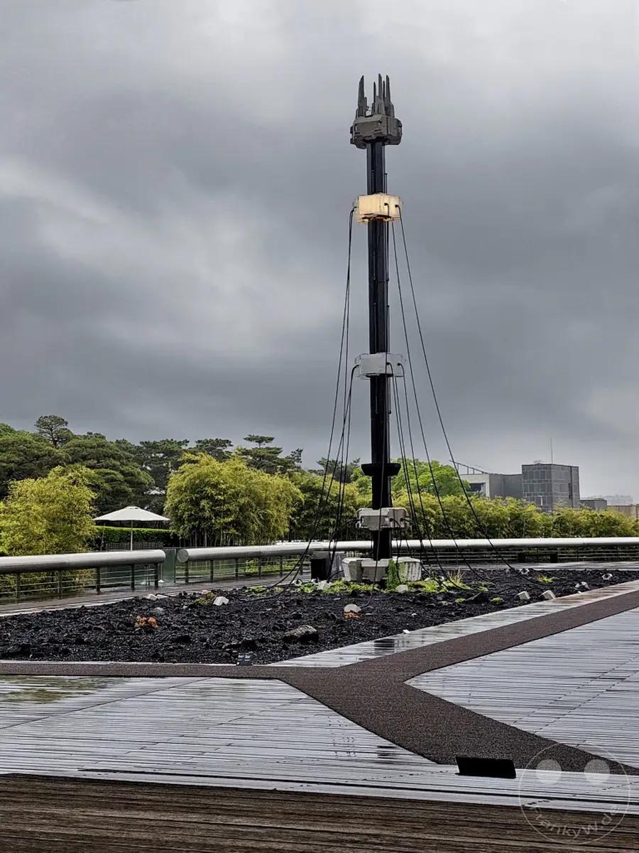 Ein markanter Skulpturenpark im Leeum Museum of Art in Seoul, Südkorea, präsentiert sich an einem bedeckten Tag. Der nasse Holzboden reflektiert das diffuse Licht, während eine hohe, säulenartige Struktur mit verschiedenen Modulen und Kabeln den Himmel durchbricht. Üppige grüne Bäume und eine Terrasse mit Sonnenschirm bilden die Kulisse für diese moderne Kunstinstallation.