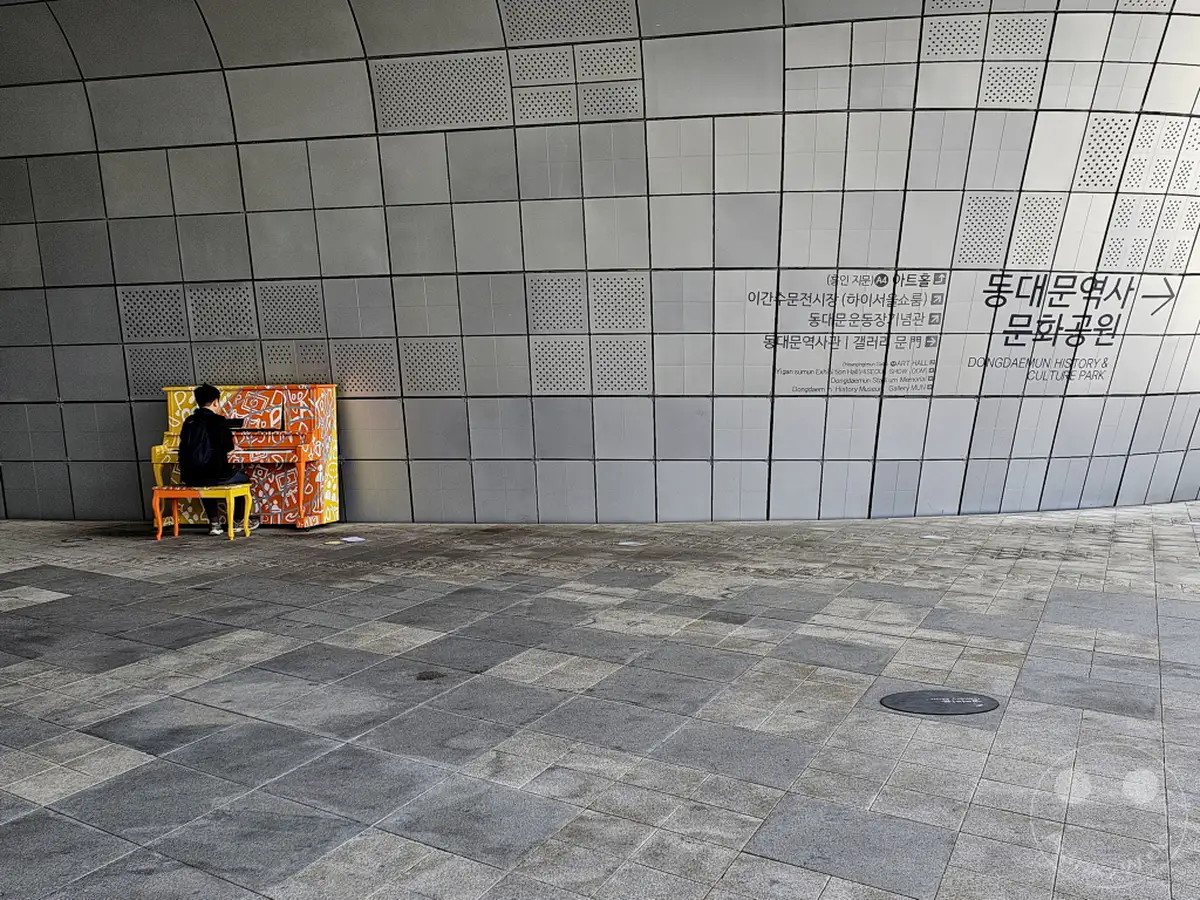 Ein einsamer Pianist spielt auf einem bunt gestalteten Klavier vor der modernen Fassade des Dongdaemun Design Plaza (DDP) in Seoul, Südkorea. Die geometrische Wand und das auffällige Kunstobjekt schaffen einen Kontrast zwischen urbaner Architektur und künstlerischem Ausdruck. Die Szene vermittelt eine ruhige und nachdenkliche Atmosphäre inmitten des städtischen Raumes.