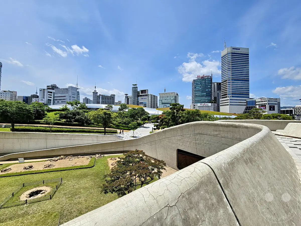 Die Skyline von Seoul, Südkorea, wird von modernen Hochhäusern dominiert, die sich hinter dem Dongdaemun Design Plaza (DDP) abzeichnen. Grünflächen und geschwungene Betonstrukturen prägen die Vordergrundansicht und schaffen einen spannenden Kontrast zwischen Natur und urbaner Architektur. Der weite, wolkenbedeckte Himmel über der Metropole versprüht eine ruhige, sommerliche Atmosphäre.