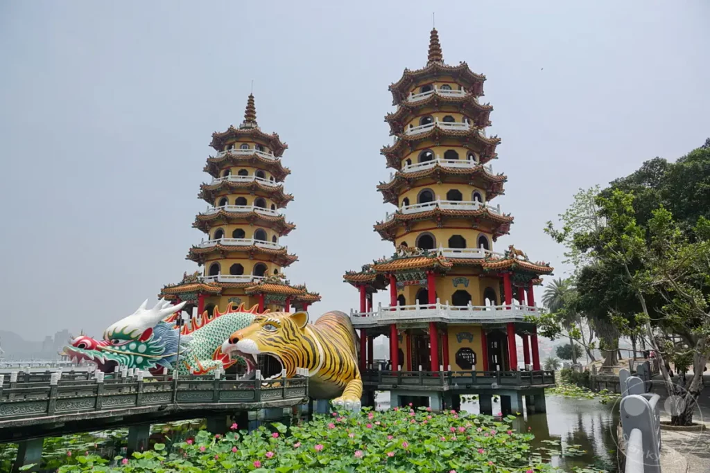 Zwei farbenprächtige, mehrstöckige Pagoden stehen auf dem Wasser in Zuoying, Taiwan, flankiert von kolossalen Skulpturen eines Tigers und eines Drachen, die den Eingang zu einer Brücke markieren. Dichte grüne Lotusblätter mit zarten rosa Blüten bedecken die Wasseroberfläche und schaffen eine ruhige und malerische Szenerie. Die Architektur und die mythologischen Kreaturen vermitteln ein Gefühl von Kultur und Spiritualität, während die üppige Natur für friedliche Atmosphäre sorgt.