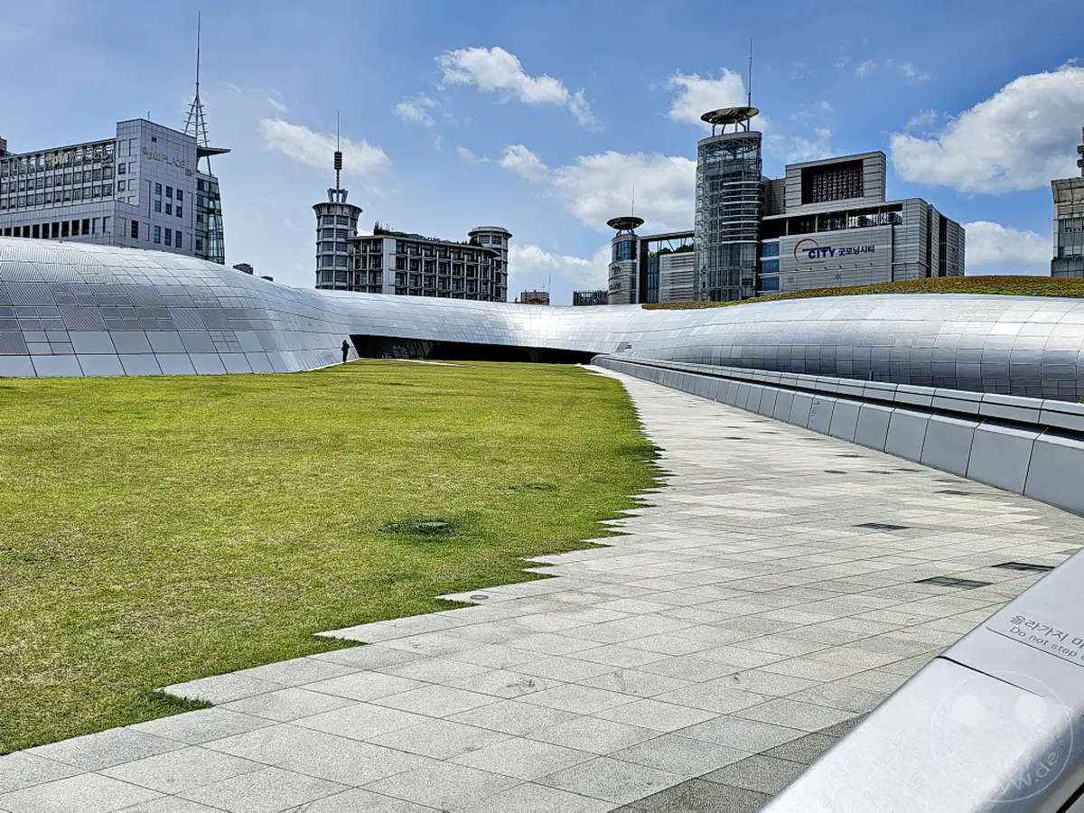 Das Dongdaemun Design Plaza (DDP) in Seoul, Südkorea, präsentiert sich mit seiner futuristischen, geschwungenen Metallfassade und den weitläufigen Grünflächen. Ein klarer Himmel mit cumulusartigen Wolken über der modernen Stadtlandschaft erzeugt eine helle und offene Atmosphäre. Die geometrischen Linien des Gehwegs und der umliegenden Gebäude bilden einen faszinierenden Kontrast zur organischen Form des DDP.