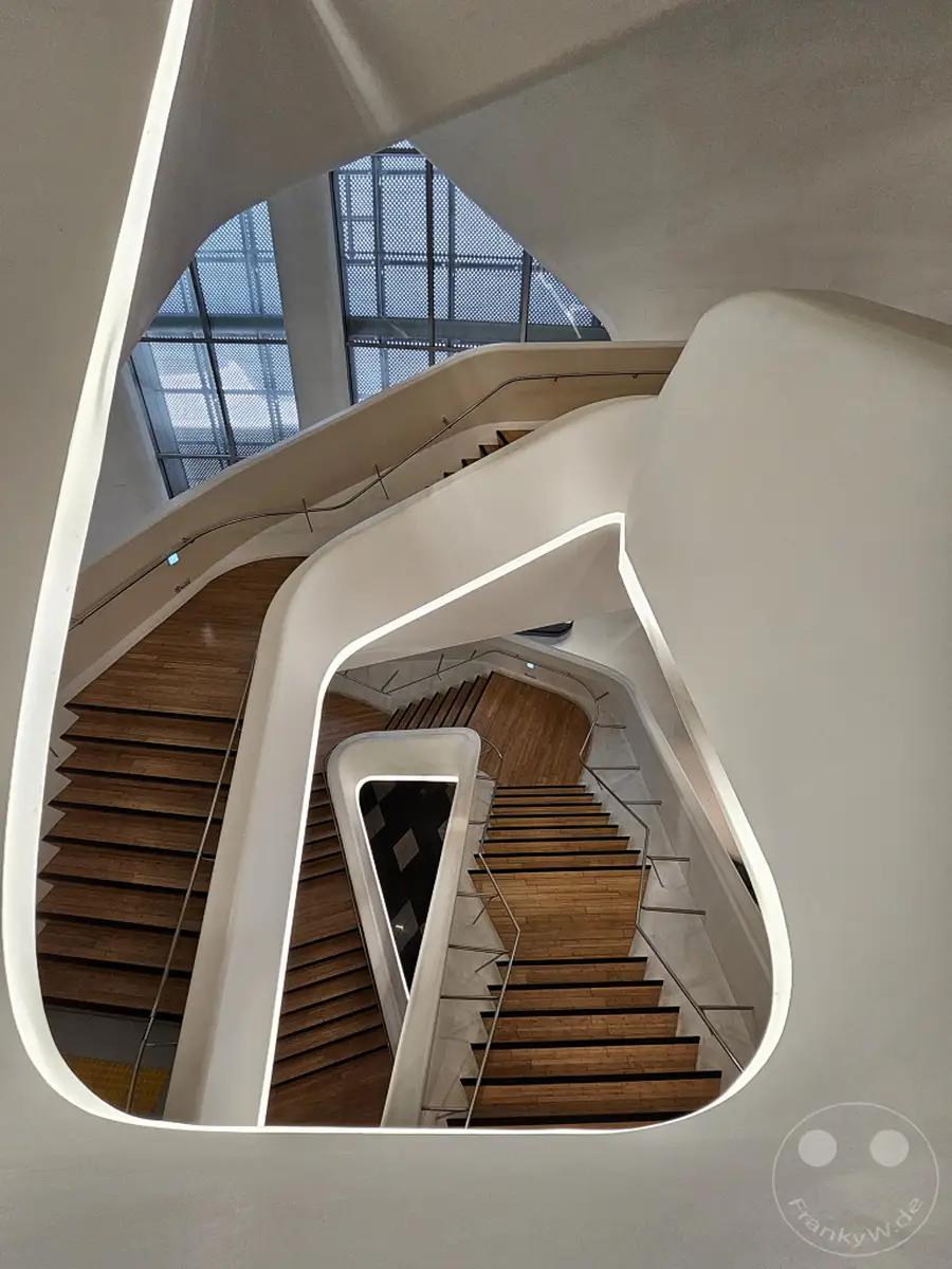Das Innere des Dongdaemun Design Plaza (DDP) in Seoul, Südkorea, präsentiert sich mit einer atemberaubenden, futuristischen Architektur. Geschwungene weiße Wände rahmen geschwungene Holztreppen ein, die sich wie ein Kunstwerk ineinander verschlingen. Die helle, kühle Beleuchtung und die klaren Linien erzeugen eine elegante und gleichzeitig dynamische Atmosphäre, die zur Erkundung einlädt.