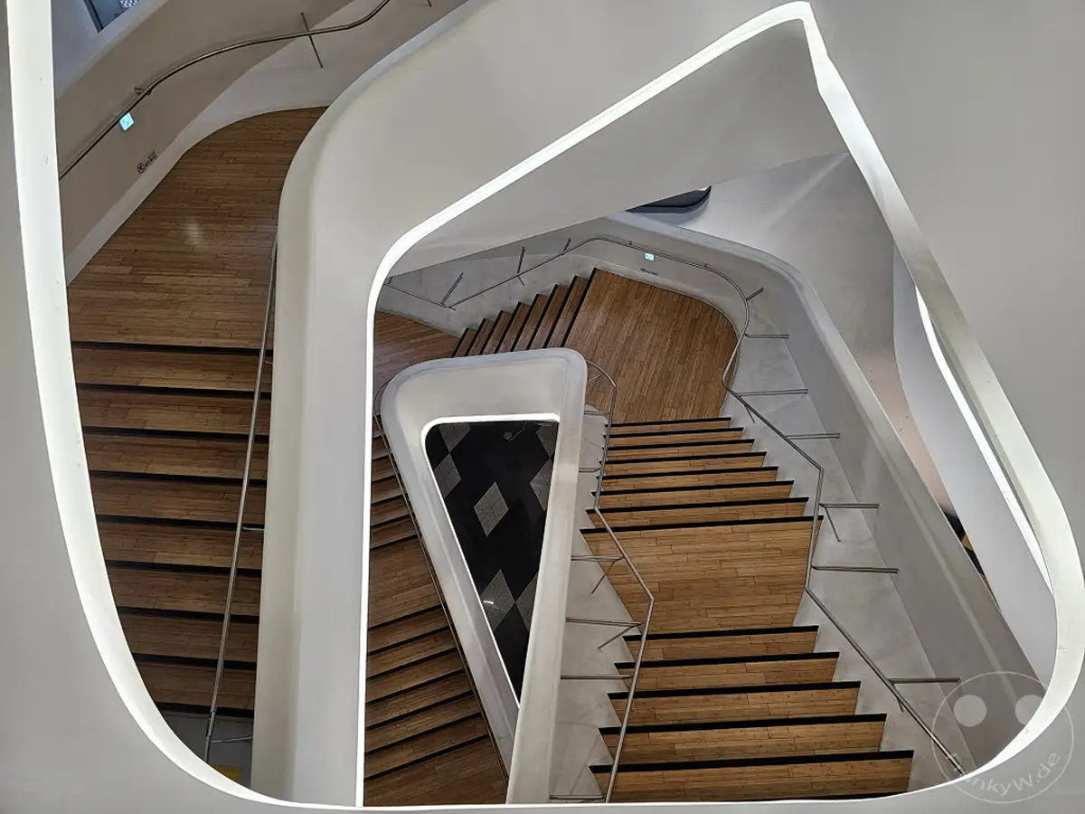 Das Innere des Dongdaemun Design Plaza (DDP) in Seoul, Südkorea, offenbart eine geschwungene, moderne Architektur mit einem beeindruckenden Treppenhaus. Helles Holz und weiße, geschwungene Elemente erzeugen eine elegante und futuristische Atmosphäre. Die Perspektive von oben betont die komplexen Linien und die Dynamik des Raumes.