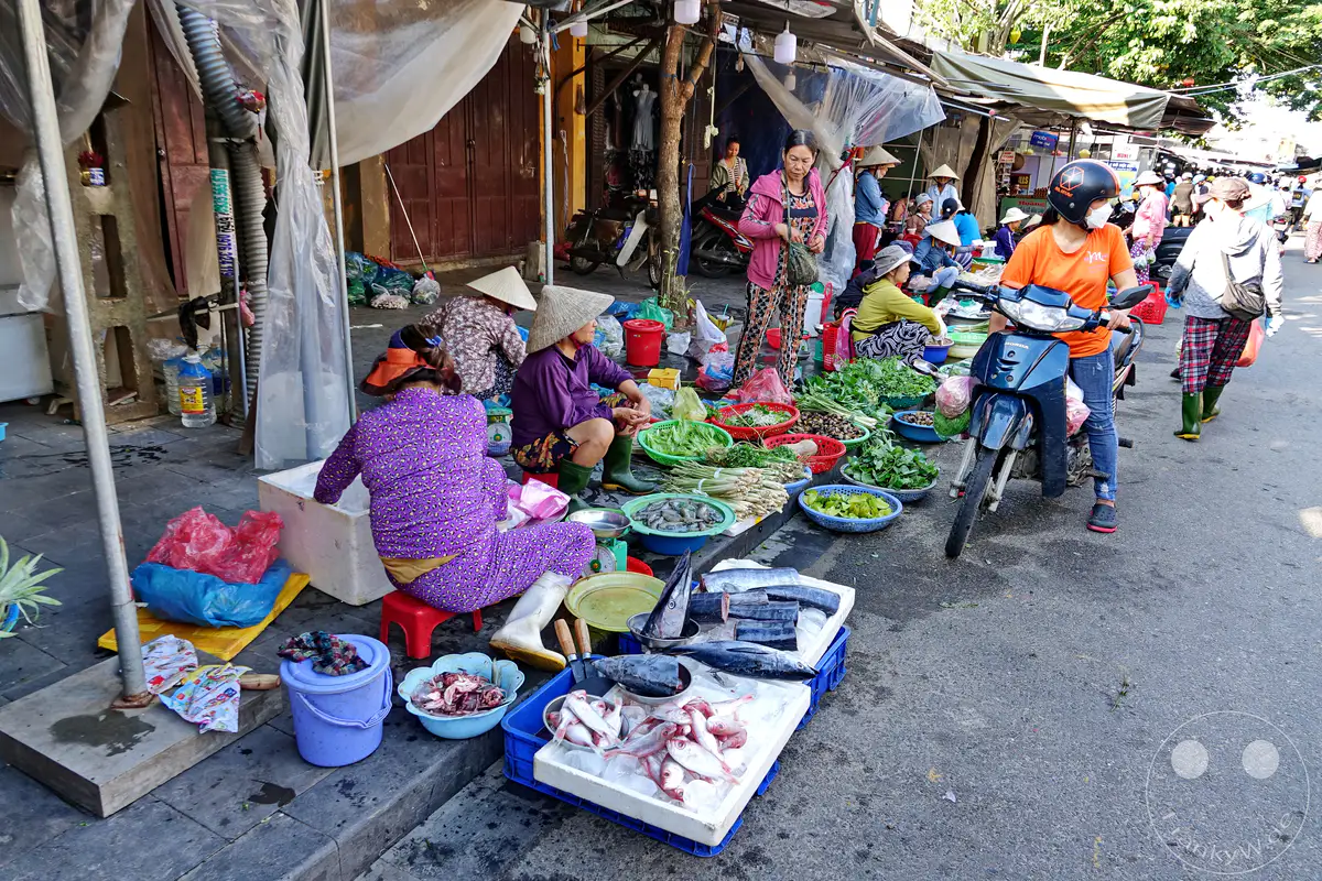 Ein lebhafter Street Market in Hoi An, Vietnam, mit einer Vielzahl von frischem Gemüse, Muscheln und Fisch, der auf dem Boden ausgebreitet und in Kisten präsentiert wird. Mehrere einheimische Frauen mit traditionellen Hüten sind damit beschäftigt, ihre Waren zu verkaufen, während Roller im Hintergrund auf der Straße parken. Die Szene vermittelt eine authentische Atmosphäre des täglichen Lebens auf einem vietnamesischen Markt.