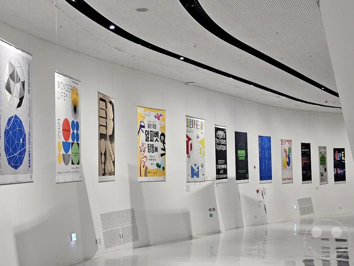 In Südkorea, Seoul, präsentiert sich der Dongdaemun Design Plaza (DDP) mit einer modernen Ausstellungsfläche. Verschiedene Banner mit grafischen Designs hängen von der Decke und säumen die geschwungene Wand. Die Szenerie vermittelt eine klare und künstlerische Atmosphäre, die typisch für Designausstellungen ist.