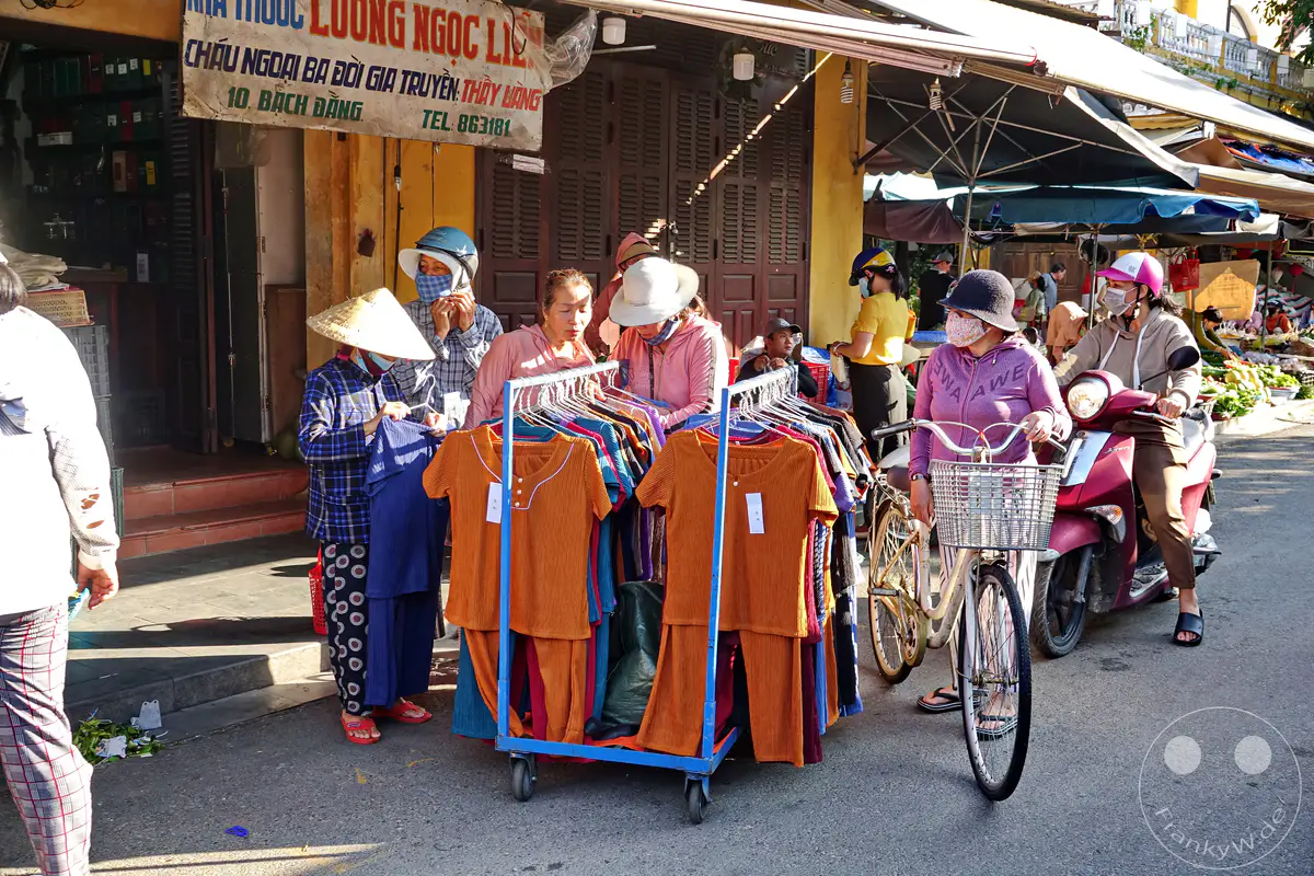 Auf einem belebten Straßenmarkt in Hoi An, Vietnam, werden bunte Kleidungsstücke auf einem Rollwagen präsentiert. Mehrere Personen, teils mit Schutzmasken und traditionellen Hüten, interagieren mit den Waren, während im Hintergrund ein Roller und weitere Marktstände zu sehen sind. Die sonnige Atmosphäre und die alltägliche Szenerie spiegeln das lebendige Treiben des Marktes wider.