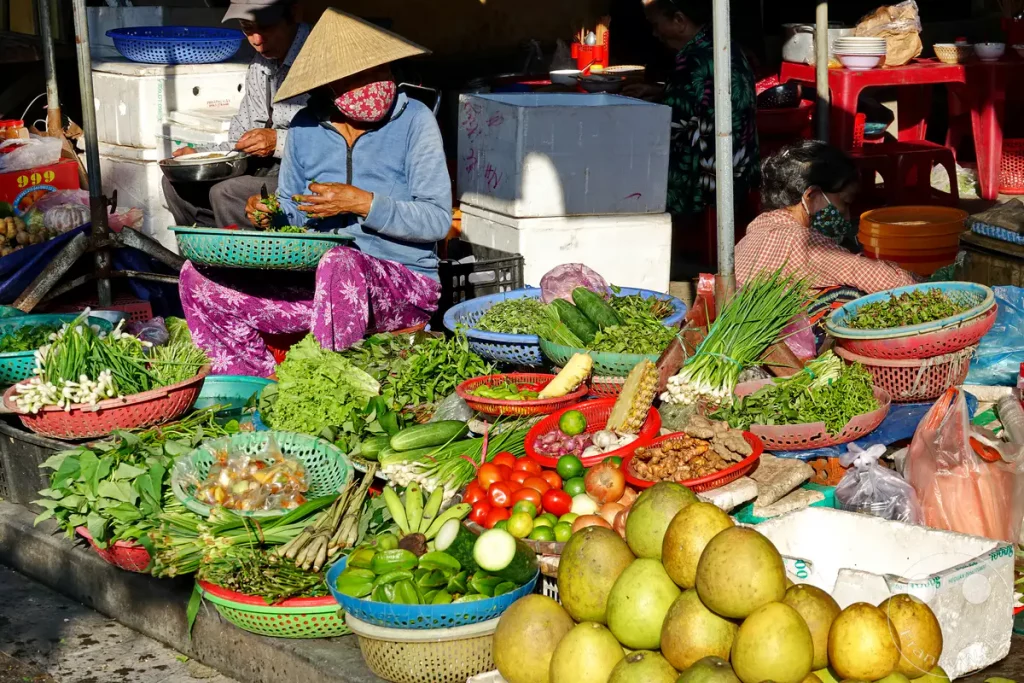 Ein geschäftiger Straßenmarkt in Hoi An, Vietnam, präsentiert eine Fülle von frischem Obst und Gemüse in bunten Körben. Ein Verkäufer mit traditionellem Hut sortiert Kräuter, während die Sonne die Szenerie beleuchtet und eine lebendige Atmosphäre schafft. Der Handel mit lokalen Produkten steht im Mittelpunkt dieses authentischen Einblicks in das tägliche Leben.
