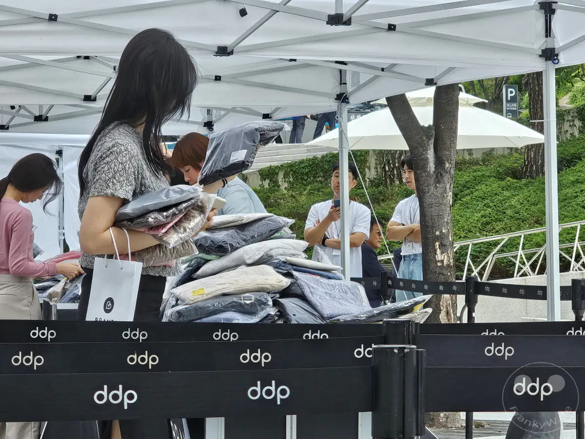 In Seoul, Südkorea, präsentieren Verkäufer auf dem Dongdaemun Design Plaza (DDP) ihre Waren, darunter auch Kleidung, die zum Verkauf angeboten wird. Die Szene erzeugt eine lebhafte und geschäftige Atmosphäre, typisch für einen Marktplatz. Eine Ausverkaufssituation wird durch die Präsentation von Kleidungsstücken angedeutet.