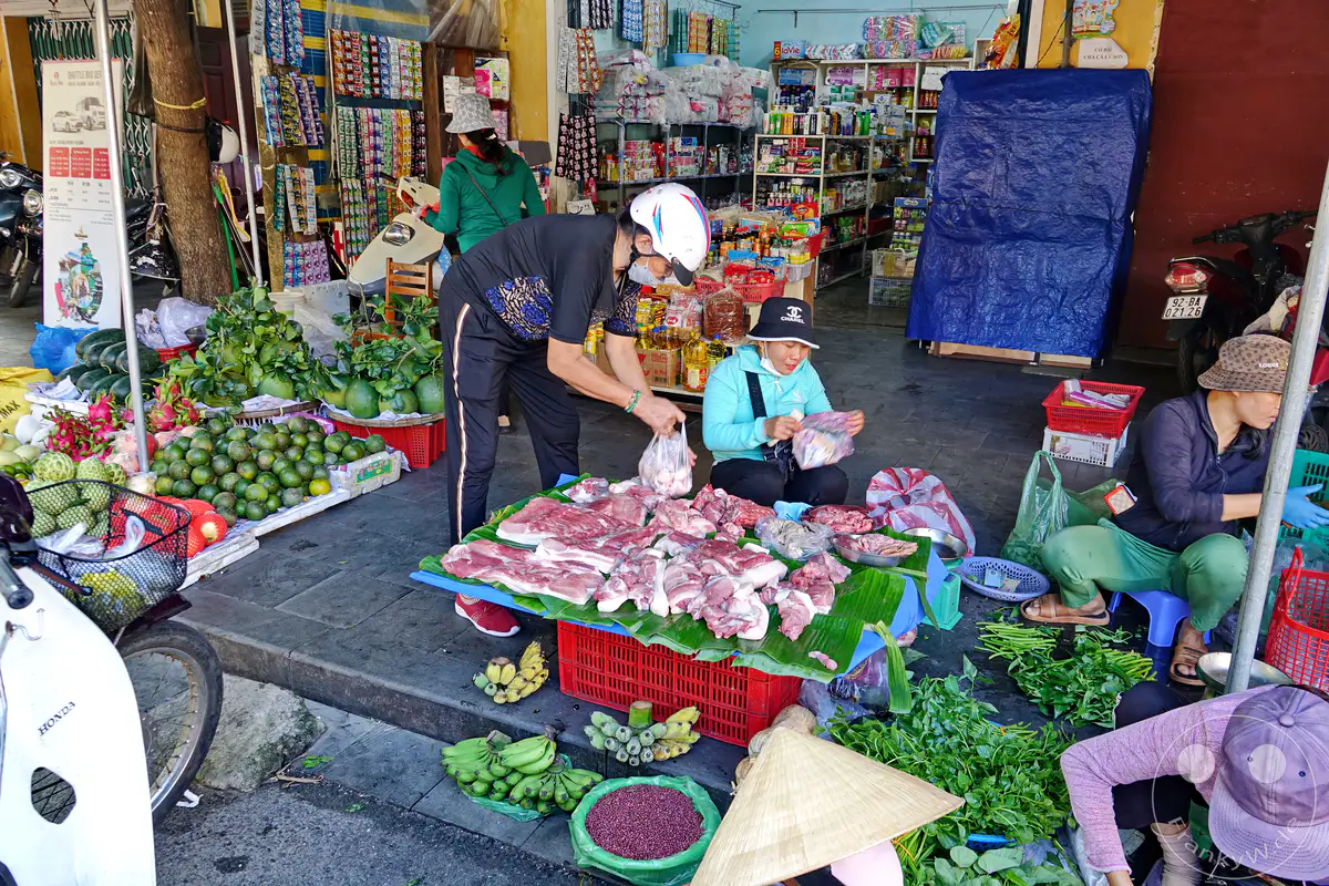 Straßenhändler in Hoi An, Vietnam, verkaufen frische Produkte und Fleisch auf einem belebten Markt. Die Szene fängt das geschäftige Treiben und die Vielfalt der angebotenen Waren ein. Kunden interagieren mit den Verkäufern inmitten von bunten Früchten, grünem Gemüse und rohem Fleisch.