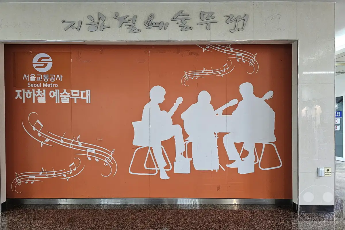 In Seoul, Südkorea, schmückt eine leuchtend orangefarbene Wand am U-Bahnhof Dongdaemun Design Plaza (DDP) eine Kunstinstallation. Silhouetten von drei Musikern, die Gitarre spielen, sind zusammen mit Notenlinien und Musiksymbolen dargestellt. Diese Szene erzeugt eine lebendige und künstlerische Atmosphäre in der U-Bahn-Station.