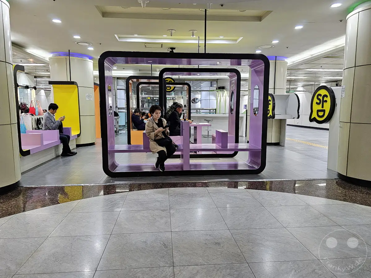 Imposante Sitzbereiche mit violetten Rahmen und Sitzflächen prägen die moderne Wartezone der Metrostation Dongdaemun Design Plaza (DDP) in Seoul, Südkorea. Reisende nutzen die bequemen Sitze, um auf ihren Zug zu warten, während sie auf ihre Smartphones schauen. Die minimalistische und funktionale Gestaltung der Station schafft eine angenehme Atmosphäre für die Pendler.