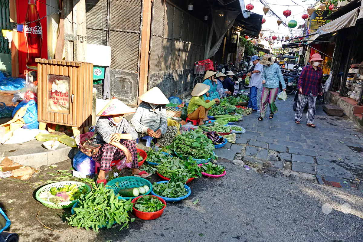 Auf einem gepflasterten Straßenmarkt in Hoi An, Vietnam, werden frische Kräuter und Gemüse zum Verkauf angeboten. Mehrere Frauen mit traditionellen kegelförmigen Hüten sitzen neben ihren Waren und bereiten diese vor. Die Szene strahlt eine authentische Alltagsatmosphäre des lokalen Handels aus.