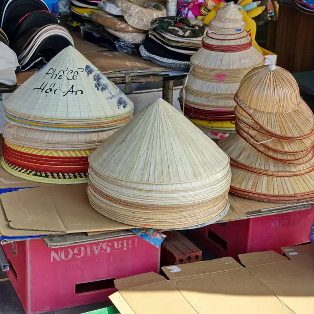 In Hoi An, Vietnam, werden auf einem Markt traditionelle kegelförmige Hüte aus Stroh zum Verkauf angeboten. Die präzise geflochtenen Hüte, teilweise mit Schriftzügen verziert, stehen stacked auf roten Kisten und Cardboard. Die Szene strahlt eine authentische Atmosphäre eines lokalen Marktes aus.