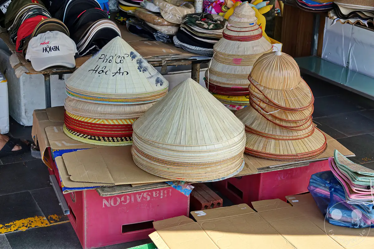In Hoi An, Vietnam, werden auf einem Markt traditionelle kegelförmige Hüte aus Stroh zum Verkauf angeboten. Die präzise geflochtenen Hüte, teilweise mit Schriftzügen verziert, stehen stacked auf roten Kisten und Cardboard. Die Szene strahlt eine authentische Atmosphäre eines lokalen Marktes aus.