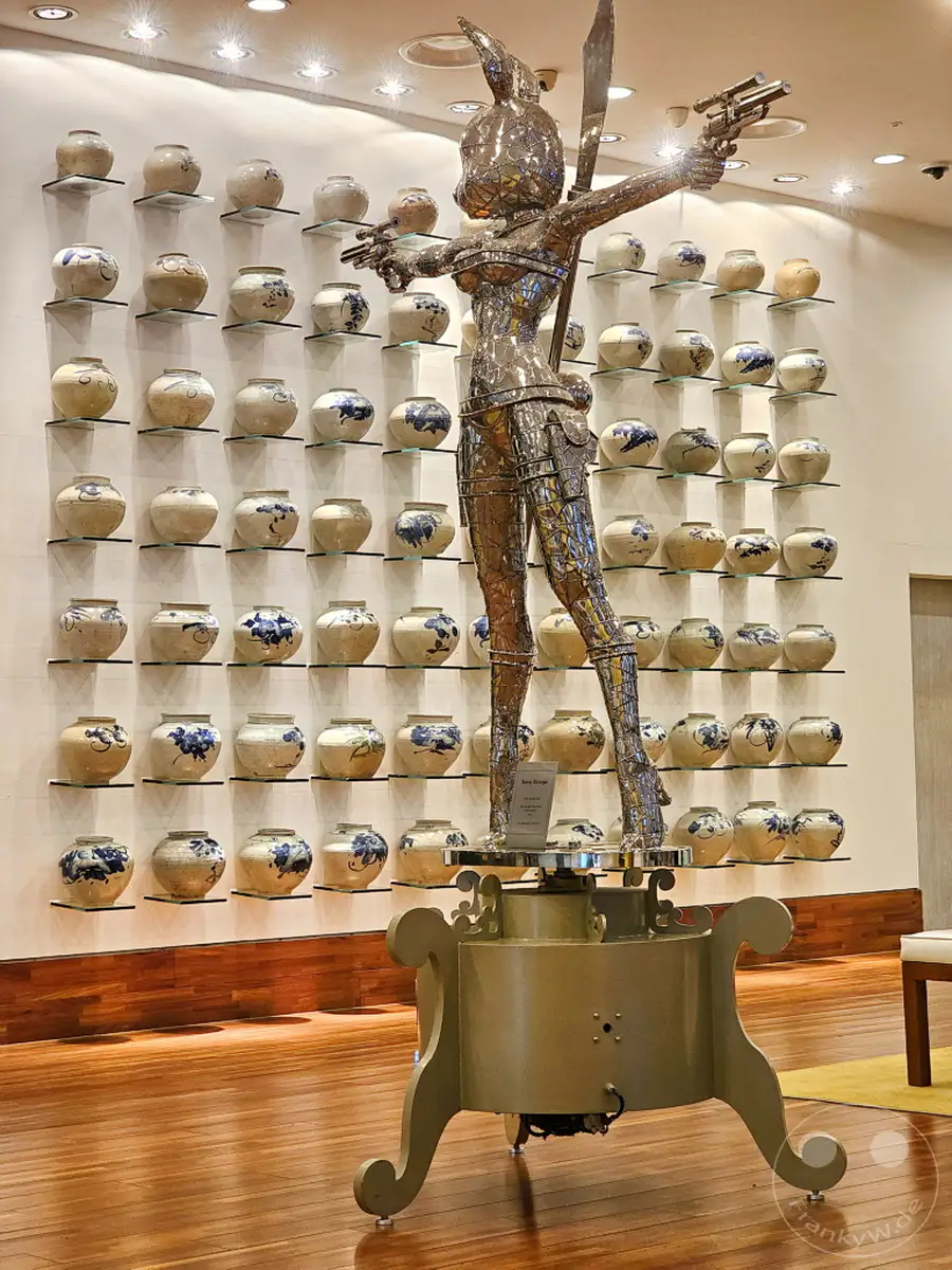 In der Lobby des Imperial Palace Boutique Hotels in Seoul steht eine auffällige Kunstinstallation einer menschlichen Figur, die wie ein Krieger mit Waffen posiert. Im Hintergrund sind an Wandregalen dutzende traditionell wirkende Keramikvasen ausgestellt, einige mit blau-weißen Mustern. Das Ambiente wirkt modern und elegant, mit einer Mischung aus zeitgenössischer Kunst und traditionellen Elementen.