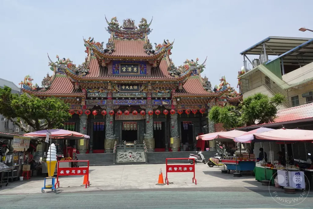 Der Chiji Tempel in Zuoying, Taiwan, präsentiert sich in voller Pracht mit seiner detailreichen Architektur und bunten Verzierungen. Vor dem Tempelgebäude herrscht ein lebhaftes Markttreiben, wo kleine Stände ihre Waren anbieten und Besucher den Ort erkunden. Die Szene vermittelt eine authentische Atmosphäre des lokalen Lebens und der religiösen Kultur.