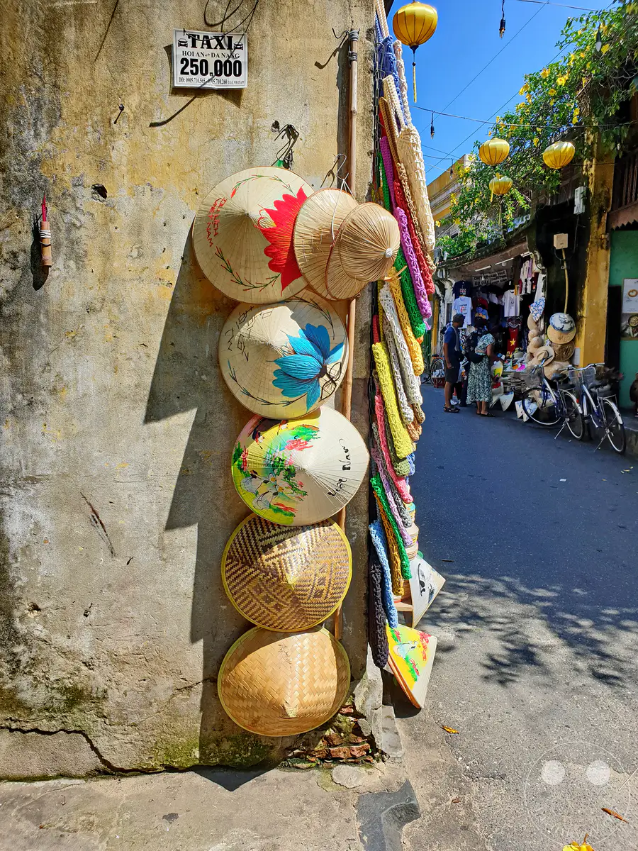 Bunte asiatische Kegelhüte, geschmückt mit traditionellen Malereien und Mustern, hängen an einer rauen, gelb verwitterten Wand in Hoi An, Vietnam. Bunte Makramee-Schnüre und gelbe Laternen säumen die Gasse und schaffen eine lebendige Atmosphäre des Markttreibens. Im Hintergrund ist ein kleiner Laden mit Kleidung und Souvenirs zu sehen, während Fahrräder am Straßenrand geparkt sind.