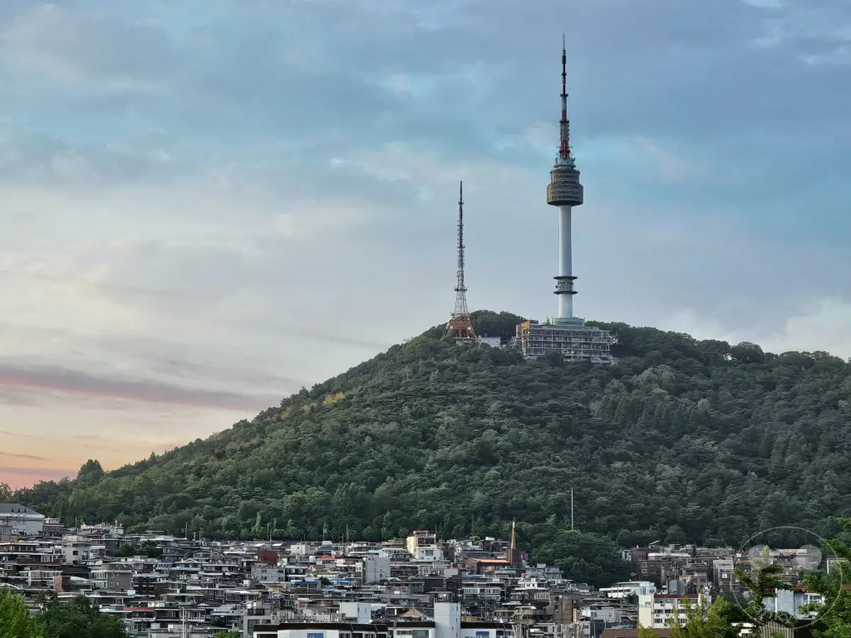 Die Silhouette von Seoul wird vom Namsan mit seinen markanten Fernsehtürmen dominiert, überragt von einer dramatischen Wolkenkulisse. Am Fuße des bewaldeten Berges erstreckt sich eine dichte Bebauung, die das städtische Leben widerspiegelt. Die Abendstimmung, eingefangen im sanften Licht, verleiht der Szene eine ruhige und zugleich majestätische Atmosphäre.