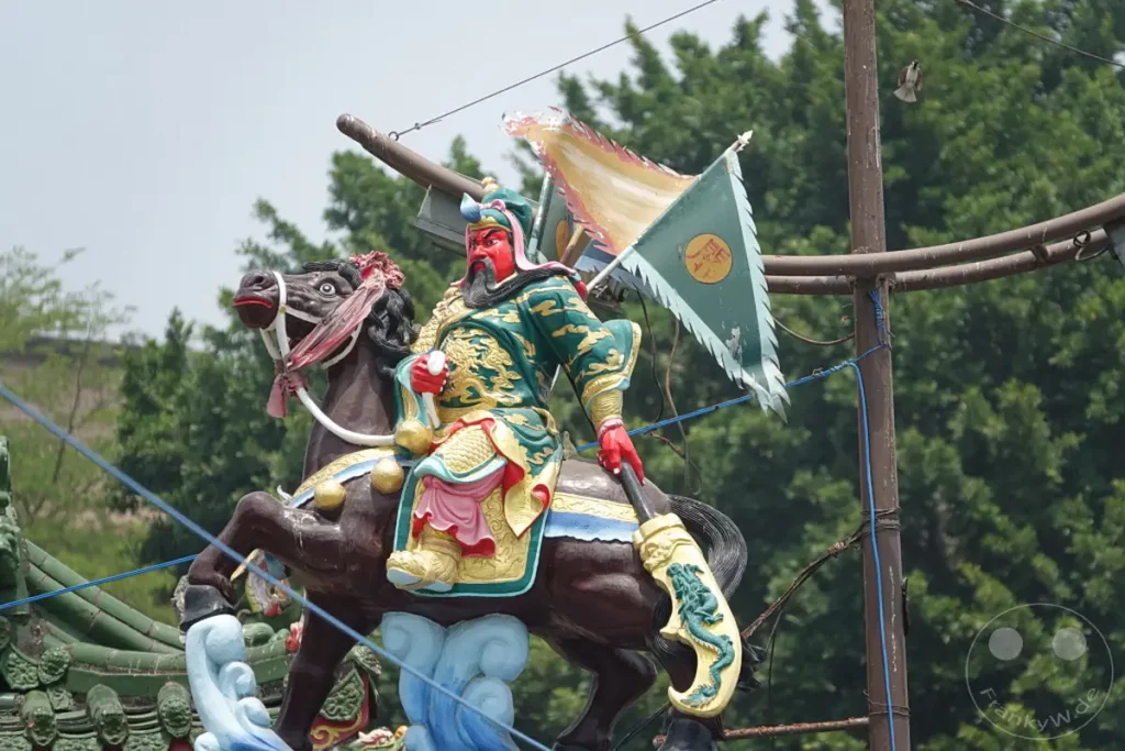 Eine bunte Skulptur eines Kriegers zu Pferd steht vor üppig grünen Bäumen in Zuoying, Taiwan. Die detailreiche Darstellung ist Teil der Spring and Autumn Pavilions und vermittelt eine feierliche und historische Atmosphäre. Das Kunstwerk ist ein eindrucksvolles Zeugnis chinesischer Kultur und Tradition.