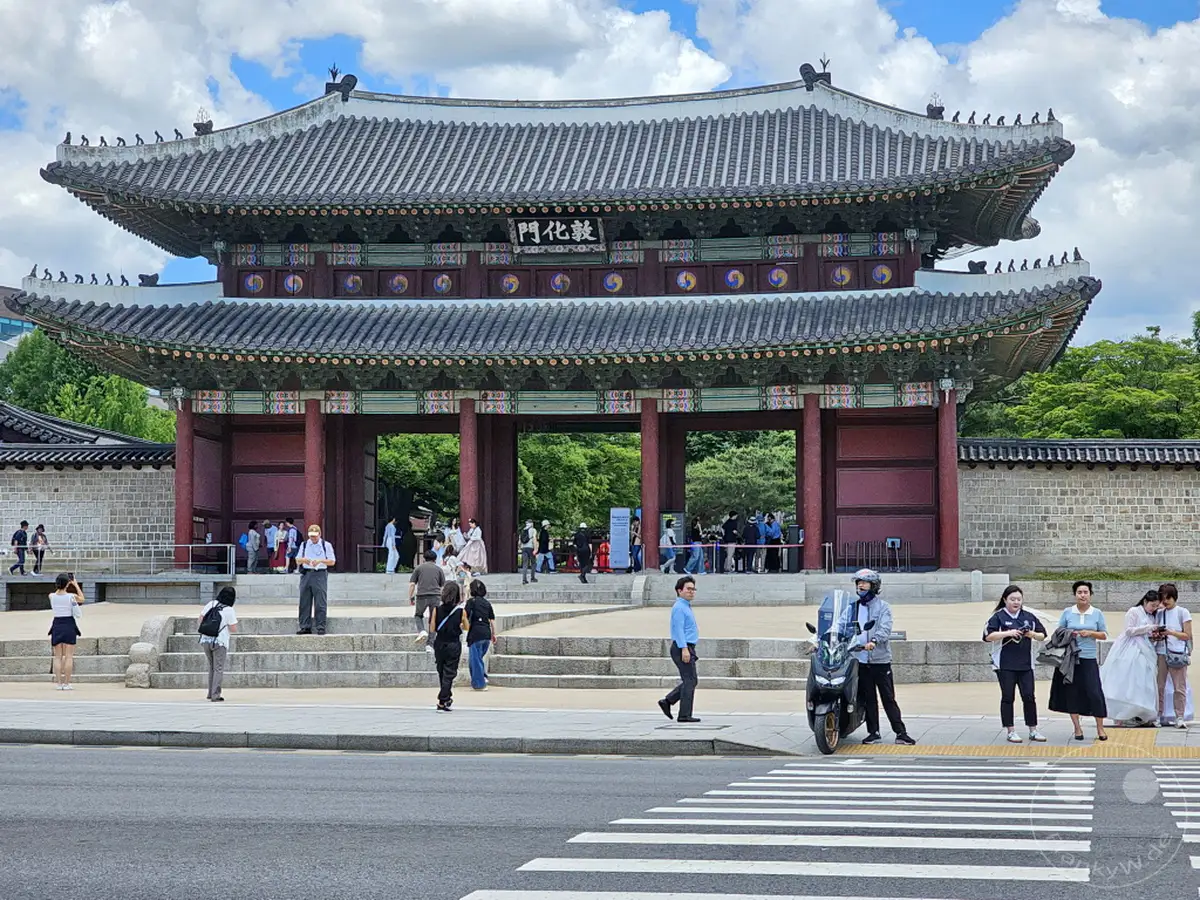 Besucher bestaunen das imposante Donhwamun-Tor des Changdeokgung-Palastes in Seoul, Südkorea. Die traditionelle Architektur und die historische Atmosphäre ziehen zahlreiche Touristen an, die die Anlage erkunden. Ein sonniger Tag mit vereinzelten Wolken bildet die Kulisse für dieses kulturelle Erlebnis.