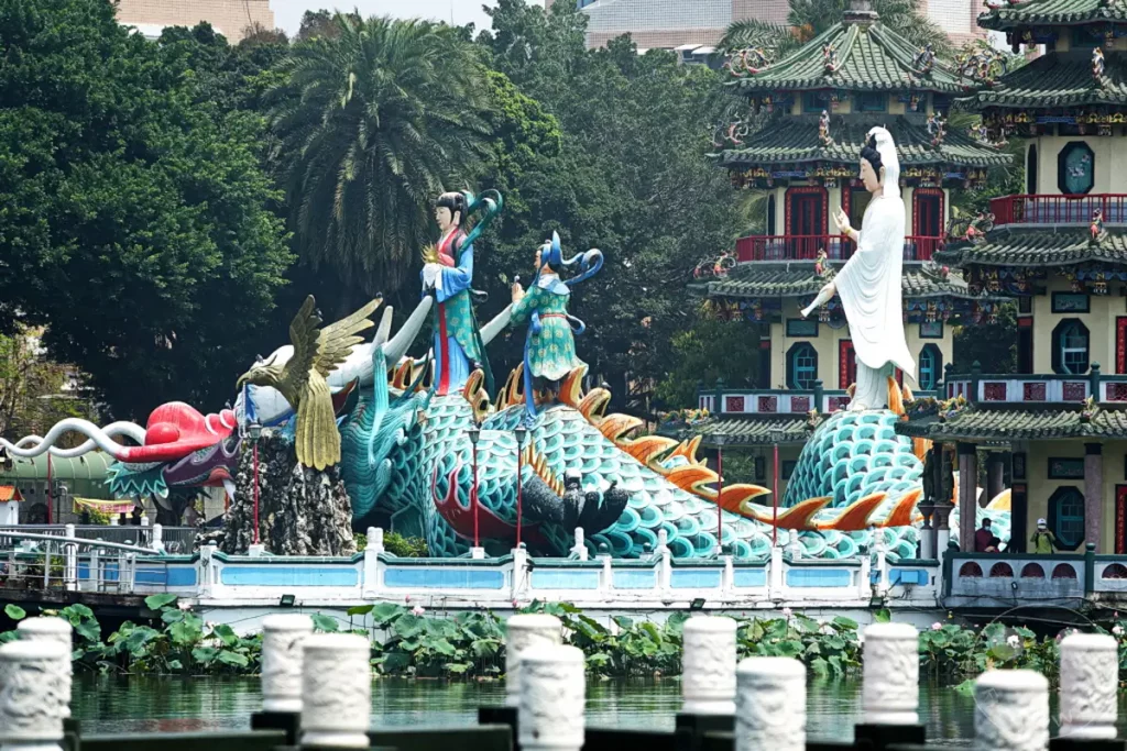 In Zuoying, Taiwan, sind kunstvolle Skulpturen eines Drachen und chinesischer Figuren vor traditionellen Tempelgebäuden platziert. Die Szenerie am Wasser mit üppigem Grün vermittelt eine ruhige und kulturell reiche Atmosphäre. Die detailreichen Darstellungen laden zu einer Entdeckungsreise in die chinesische Mythologie ein.