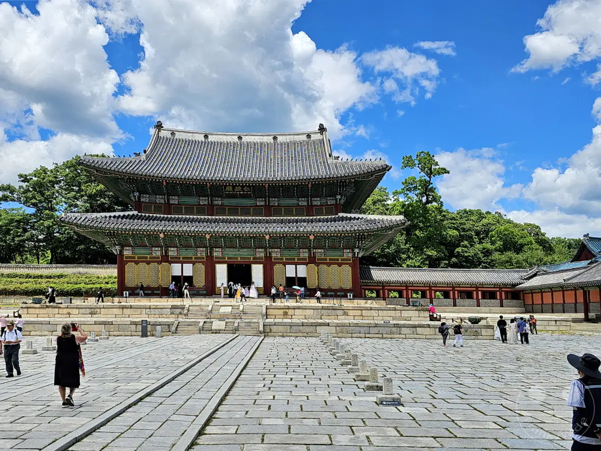 Der Changdeokgung Palast in Seoul, Südkorea, präsentiert sich an einem sonnigen Tag unter einem blauen Himmel mit weißen Wolken. Besucher schlendern über den weitläufigen, mit Steinplatten ausgelegten Hof und bewundern die kunstvolle traditionelle koreanische Architektur des Palastes. Die Szene strahlt eine Atmosphäre der Ruhe und historischen Erhabenheit aus.