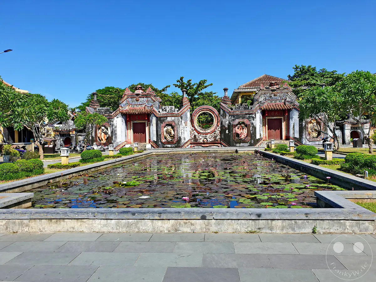 Der Ba Mu Tempel in Hoi An, Vietnam, präsentiert sich mit dem imposanten Tam Quan Tor, das von einem Teich mit zahlreichen Lotusblumen umgeben ist. Die traditionelle Architektur spiegelt die reiche Geschichte und Kultur der Region wider und vermittelt eine friedliche und zeitlose Atmosphäre. Die klare blaue Himmelkulisse bildet einen eindrucksvollen Kontrast zu den kunstvoll verzierten Gebäuden und dem üppigen Grün.
