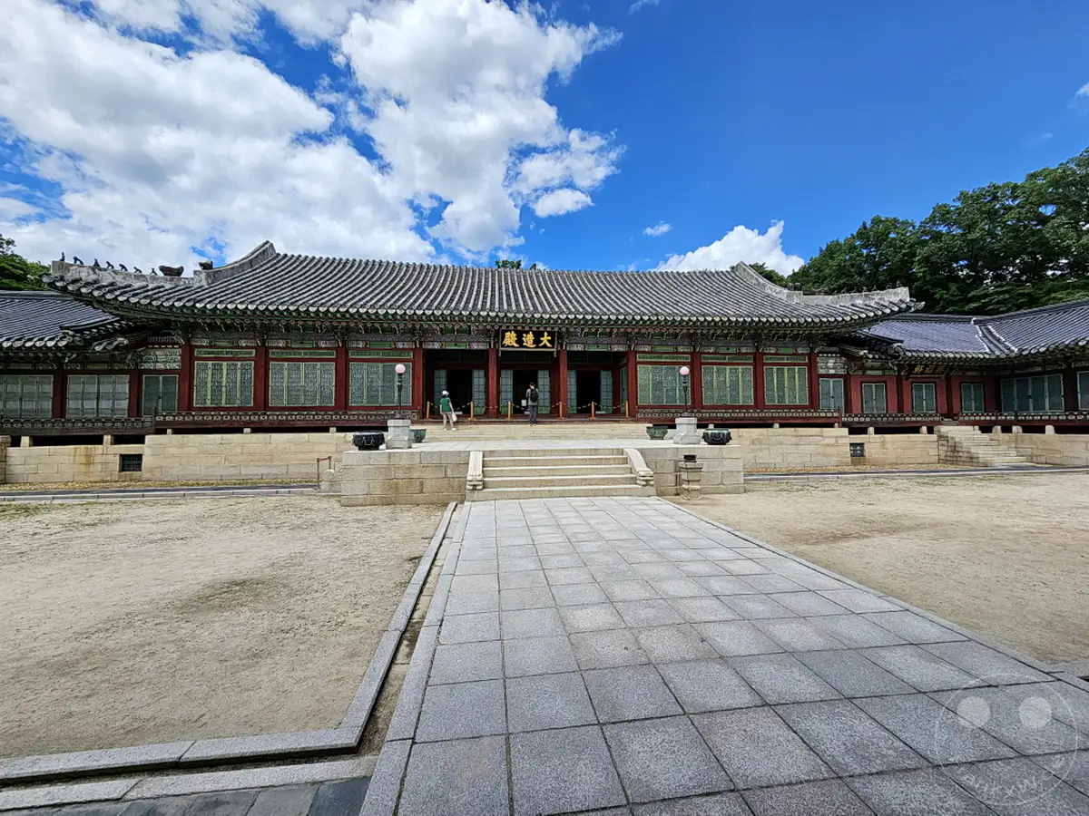 Der Changdeokgung Palast in Seoul, Südkorea, präsentiert sich unter einem strahlend blauen Himmel mit dramatischen Wolkenformationen. Die traditionelle koreanische Architektur mit ihren geschwungenen Dächern und roten Akzenten bildet einen eindrucksvollen Kontrast zur natürlichen Umgebung. Die Szene strahlt eine Atmosphäre von historischer Erhabenheit und friedlicher Schönheit aus.