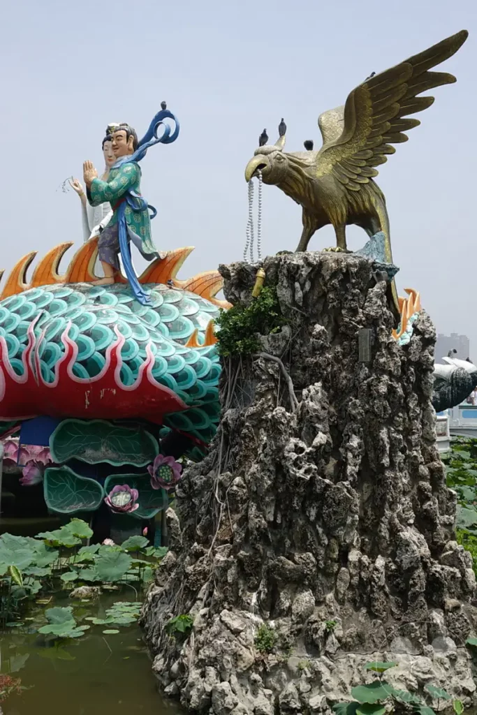Das Bild zeigt die beeindruckenden Spring and Autumn Pavilions in Zuoying, Taiwan. Eine Drachenbootskulptur und eine Skulptur eines Greifvogels dominieren die Szenerie, umgeben von Lotusblumen und einem Teich. Die detaillierten Kunstwerke und die üppige Vegetation schaffen eine faszinierende und mystische Atmosphäre.