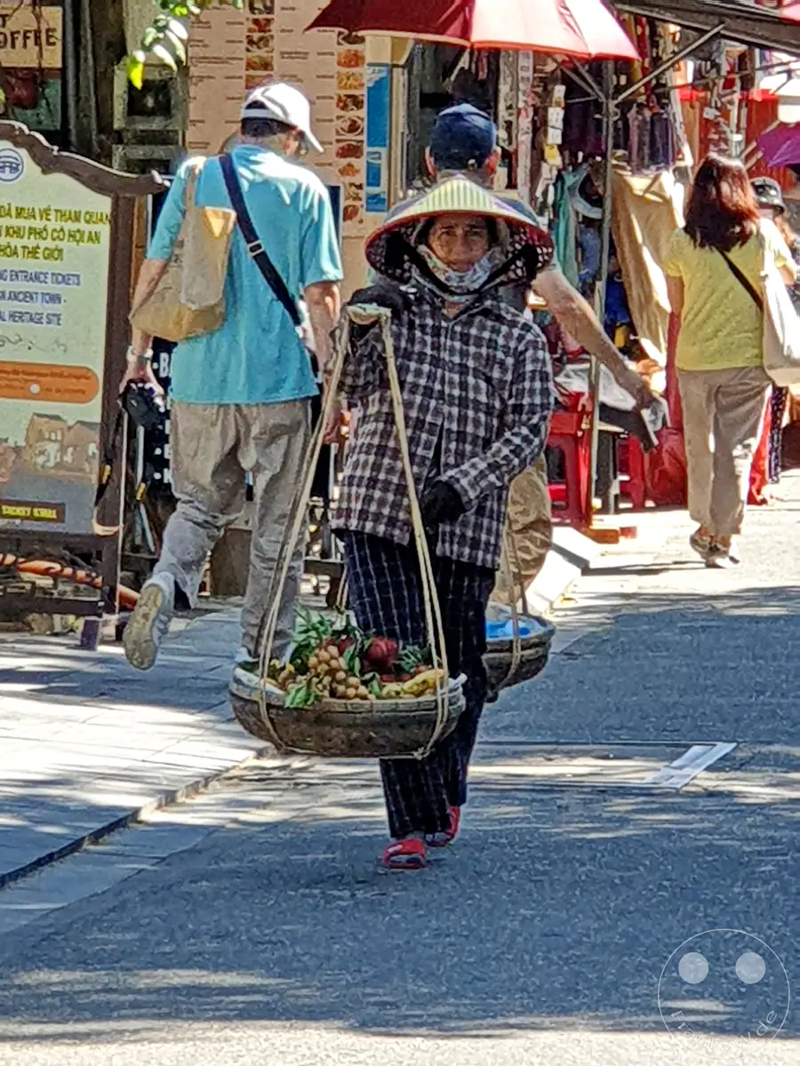 Eine Verkäuferin trägt mit einem Joch einen Korb voller bunter Früchte durch die Gassen von Hoi An, Vietnam. Menschen schlendern im Hintergrund an Marktständen vorbei, während die Sonne den Boden in ein Spiel aus Licht und Schatten taucht. Die Szene fängt das lebendige Treiben und die traditionelle Atmosphäre eines asiatischen Marktes ein.