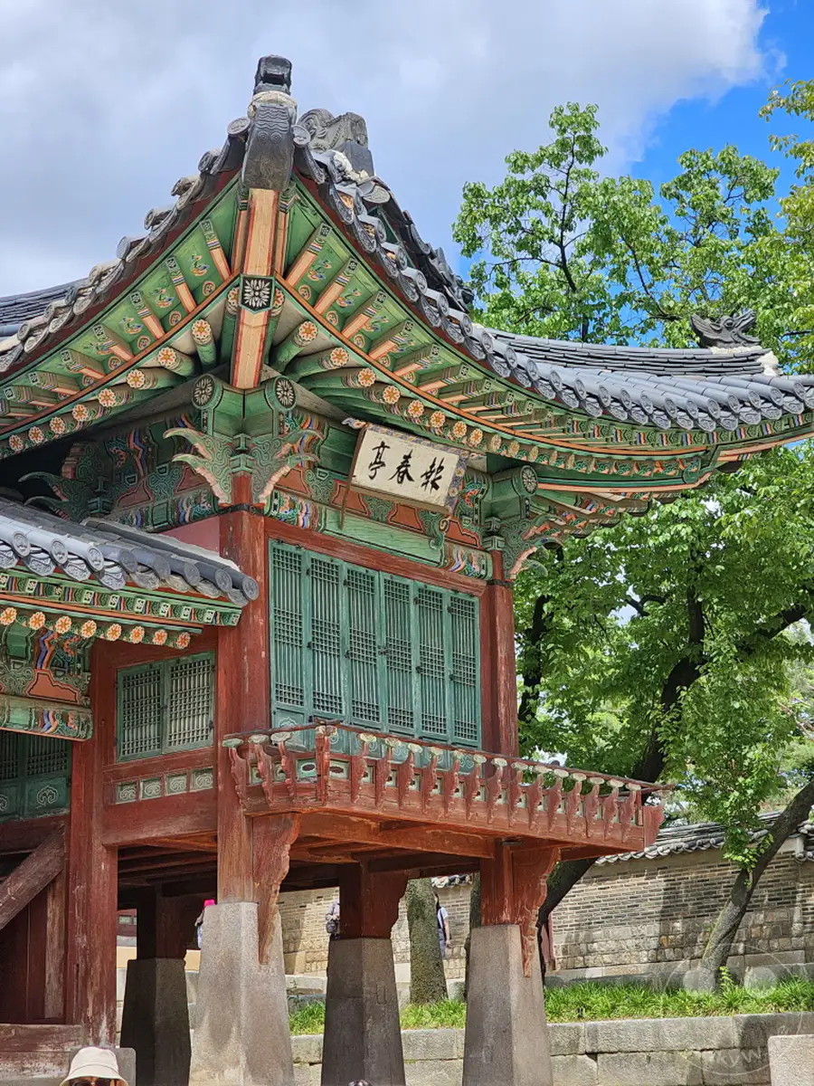 Ein detailreicher Blick auf die traditionelle Architektur des Changdeokgung Palastes in Seoul, Südkorea. Die kunstvollen Bemalungen auf den Balken und die filigranen grünen Fensterläden zeugen von meisterhafter Handwerkskunst. Im Hintergrund ragen alte Bäume unter einem blauen Himmel mit weißen Wolken auf und verleihen der Szene eine friedliche und historische Atmosphäre.