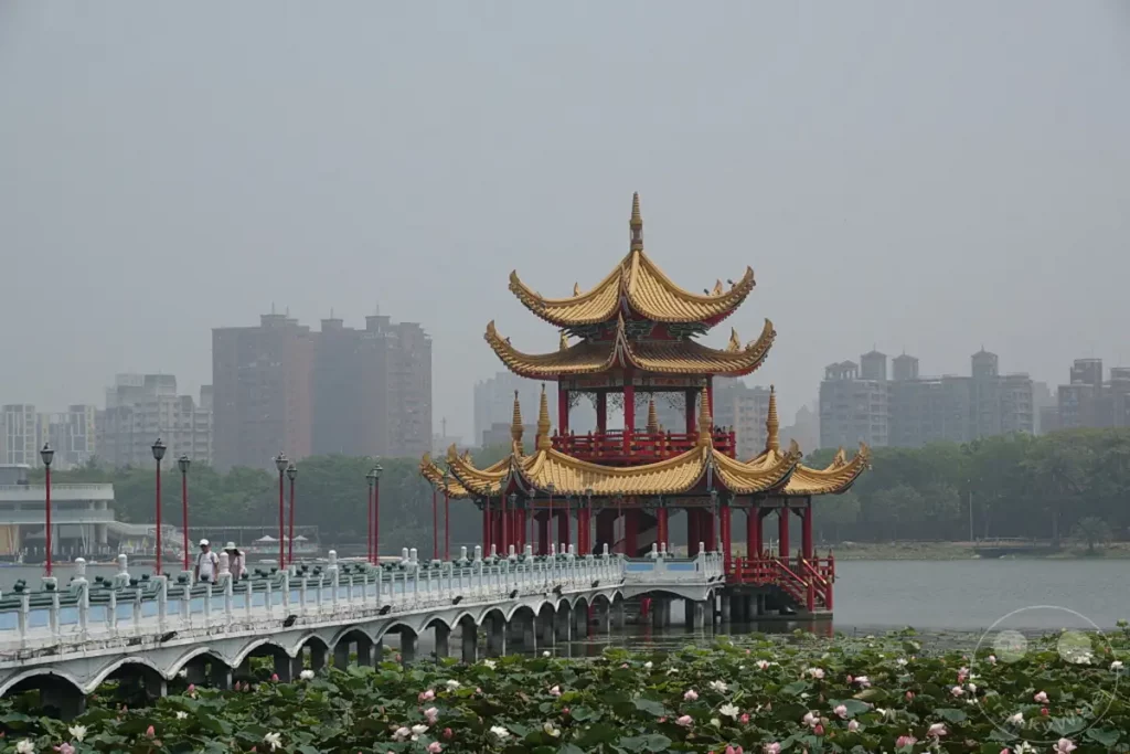 Eine malerische Szene in Zuoying, Taiwan, präsentiert die ikonische Wuliting-Pagode, die auf dem Wasser thront, umgeben von üppigen Lotusblüten und spiegelnden Wasserflächen. Im Hintergrund erhebt sich eine moderne Stadtkulisse über grünen Baumwipfeln, was einen faszinierenden Kontrast zwischen Tradition und urbanem Leben schafft. Die Atmosphäre ist ruhig und friedlich, unterbrochen nur vom sanften Rauschen des Wassers und dem Geplauder der wenigen Spaziergänger auf der Brücke.
