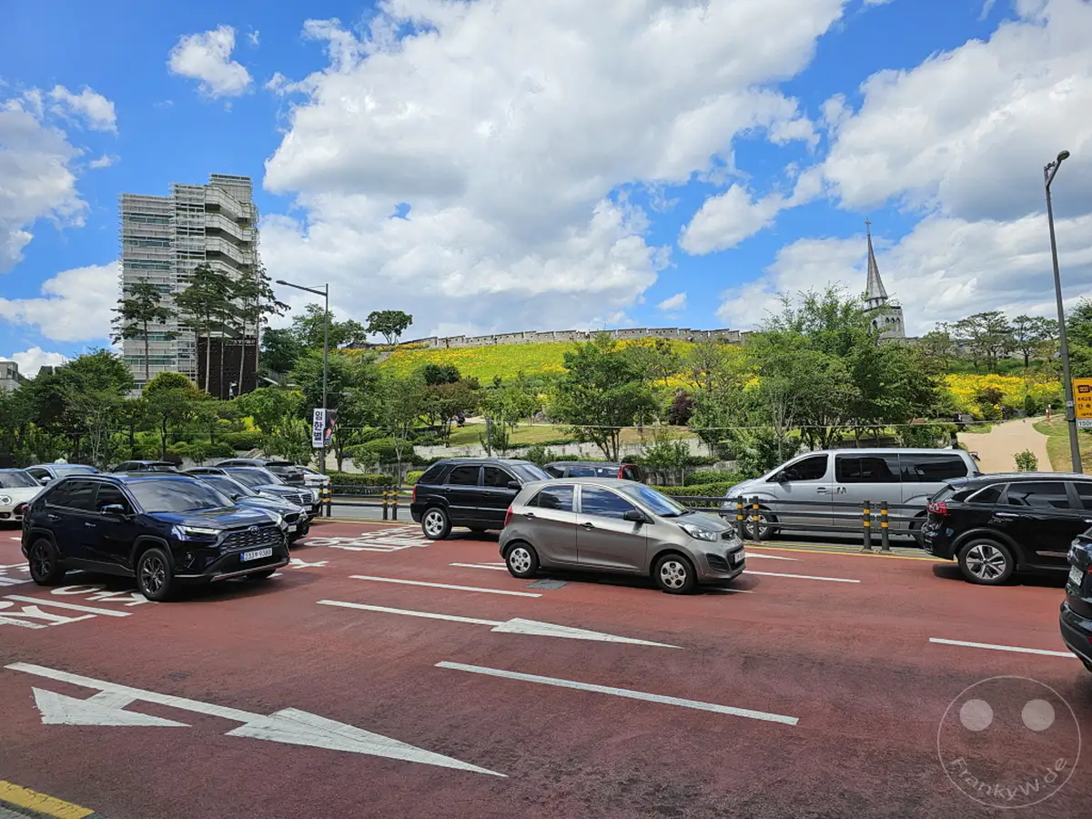 Ein sonniger Tag in Seoul, Südkorea, mit dem imposanten Hwaseong-Festungsmauer im Hintergrund. Autos bewegen sich auf einer roten Fahrbahn mit weißen Pfeilen, während am Horizont eine moderne Hochhausarchitektur und die historische Stadtmauer aufeinandertreffen. Der Himmel ist mit dramatischen Wolkenformationen geschmückt, die der Szene eine lebendige und dynamische Atmosphäre verleihen.
