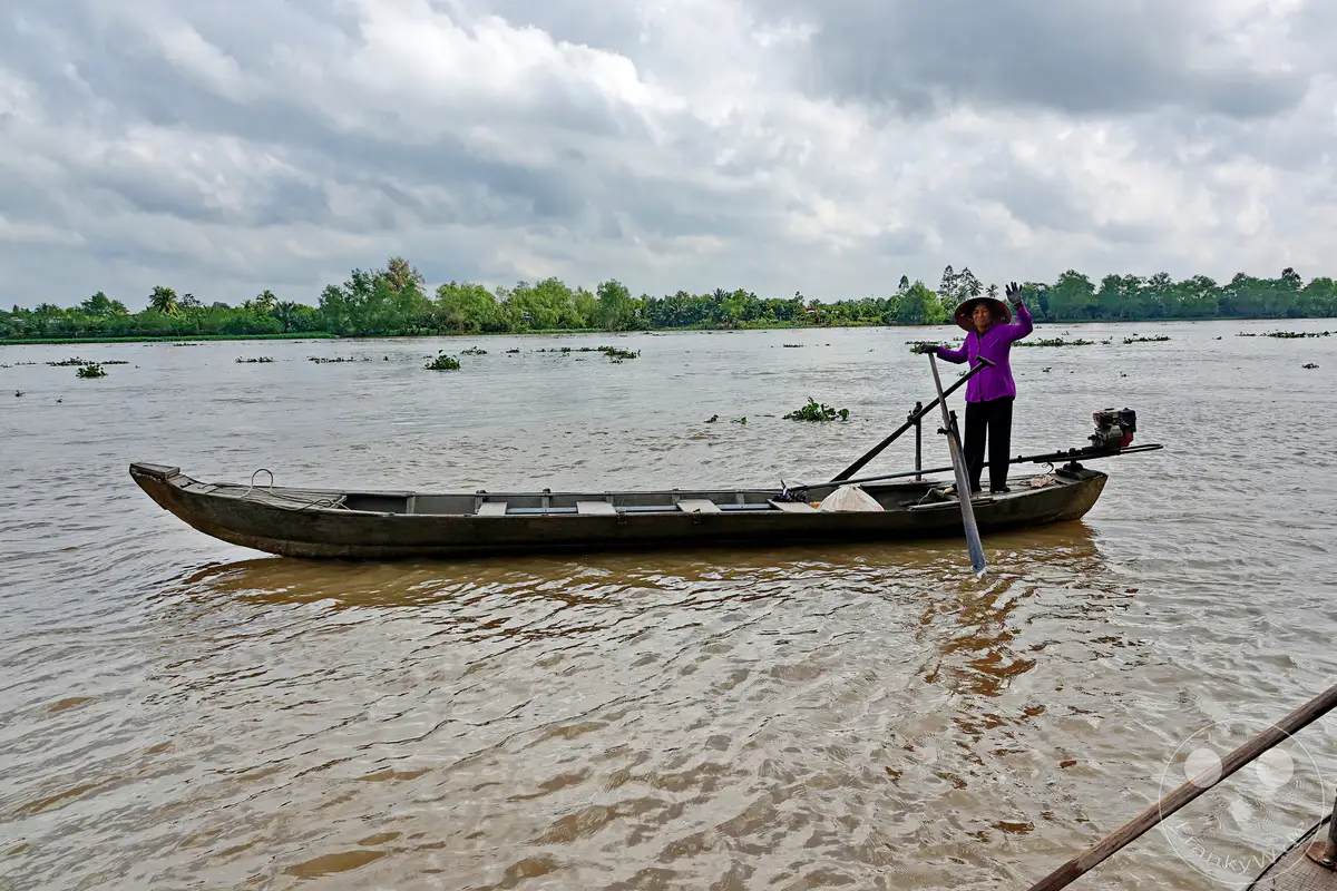 Eine Frau in traditioneller Kleidung und Hut steuert ein Holzboot auf dem trüben Mekong in Vietnam. Sie hält einen Ruderpaddel in der Hand und hebt eine winkende Hand, während sie auf dem Wasser unterwegs ist. Die üppige grüne Ufervegetation und der bewölkte Himmel bilden die Kulisse für diese ruhige Szene auf dem Fluss.