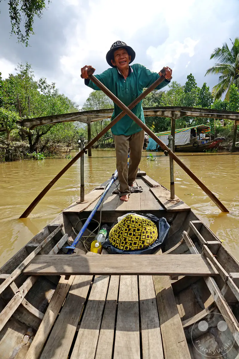 Ein älterer Mann steuert mit zwei Rudern ein Holzboot auf einem schlammigen Fluss in Vietnam. Die üppige grüne Uferlandschaft und der bewölkte Himmel schaffen eine ruhige, aber lebendige Atmosphäre. Im Vordergrund liegt ein bunt gemustertes Sitzkissen in einem schwarzen Plastiksack neben einer Wasserflasche.