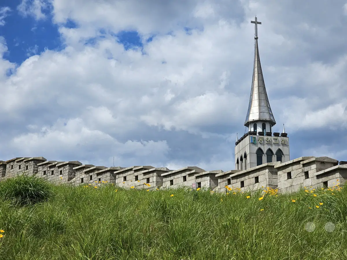 Die imposante Festungsmauer von Seoul erhebt sich über einem üppigen grünen Hügel, der mit gelben Wildblumen übersät ist. Über der historischen Struktur thront eine moderne Kirche mit einem schlanken Turm und einem Kreuz, das sich gegen einen dramatisch bewölkten Himmel abzeichnet. Diese Szene in Seoul, Südkorea, vereint Geschichte, Natur und Spiritualität in einer malerischen Kulisse.