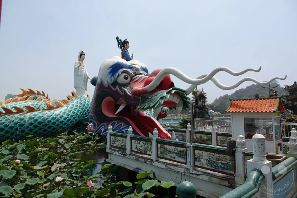 In Taiwan, Zuoying, am Ufer des Sees, thront eine beeindruckende Skulptur eines bunten Drachenkopfes mit aufgesperrtem Maul, aus dem eine rote Rutsche führt. Im Hintergrund erhebt sich die majestätische Statue einer weiblichen Gottheit vor einem klaren Himmel. Die Szenerie wird durch einen ruhigen Teich, bedeckt mit grünen Lotusblättern und zarten rosa Blüten, belebt, was eine friedliche und kulturell reiche Atmosphäre schafft.