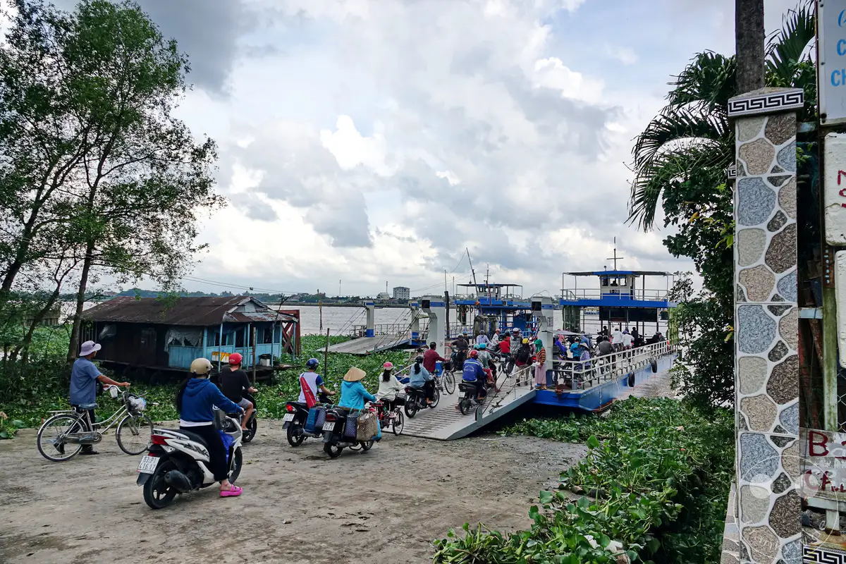 In Vietnam ist eine Fähre voller Motorroller und Menschen auf dem Mekong unterwegs. Dichtes grünes Wassergras säumt das Ufer, während die Wolken einen dramatischen Himmel malen. Die Szene strahlt geschäftige Betriebsamkeit inmitten einer tropischen Flusslandschaft aus.