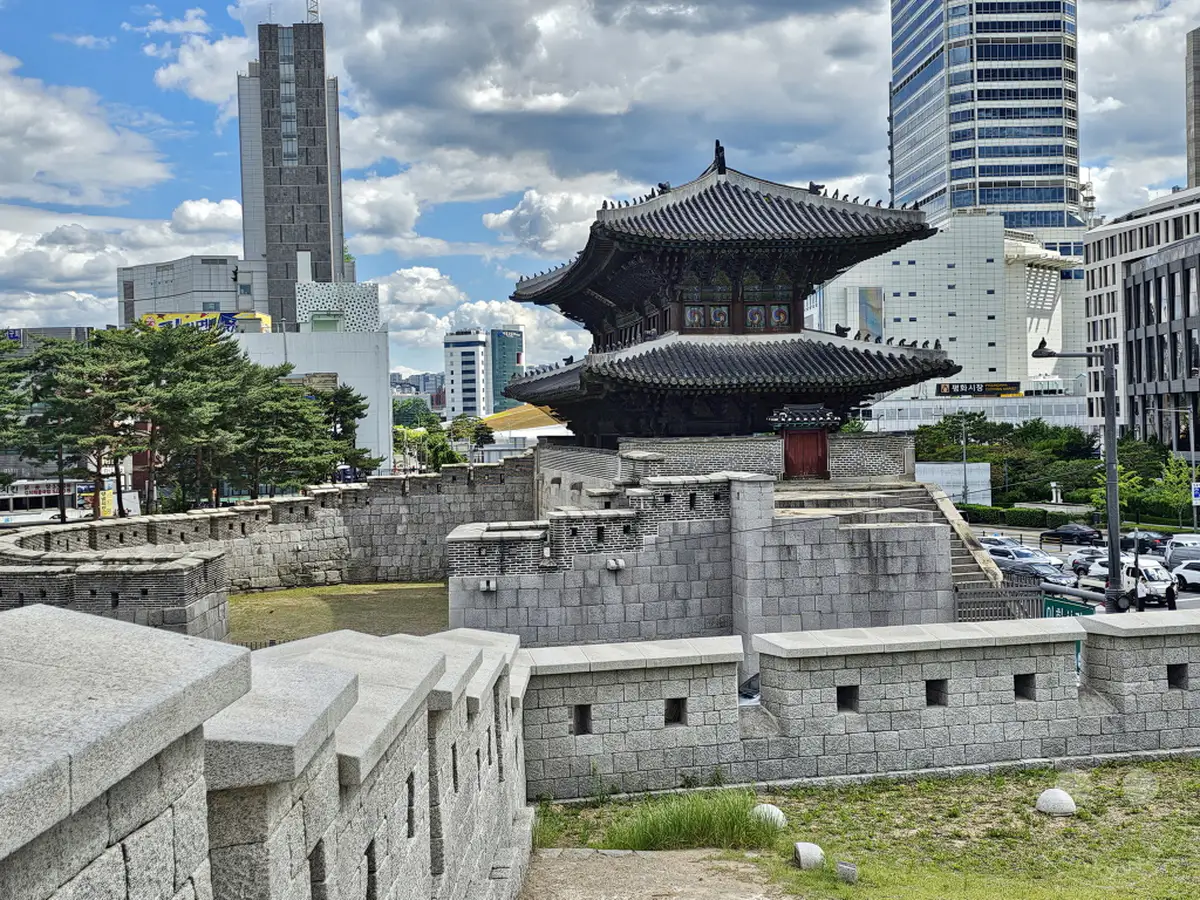 Eine historische Festungsmauer, die sich durch die geschäftige Metropole Seoul schlängelt, steht im Kontrast zu modernen Hochhäusern am Himmel. Die traditionelle Architektur der Festung mit ihren steinernen Mauern und dem verzierten Dach eines Gebäudes inmitten des Komplexes strahlt Ruhe aus. Die Szene vermittelt eine faszinierende Mischung aus alter Geschichte und pulsierender Gegenwart in der Hauptstadt Südkoreas.