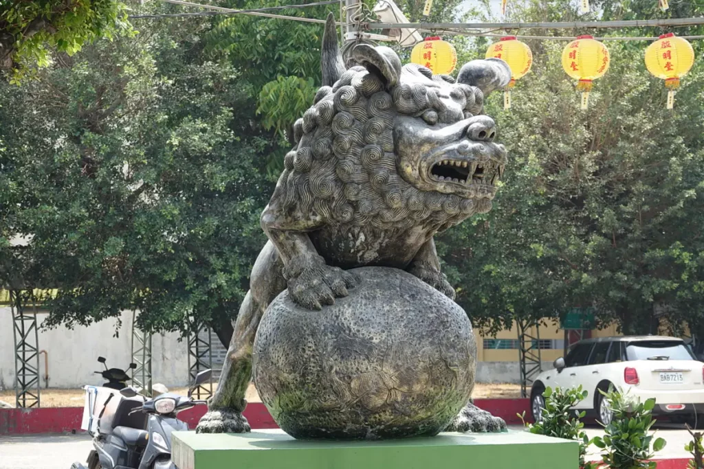 Vor dem Chiji Tempel in Zuoying, Taiwan, thront eine detailreiche Löwenstatue aus Stein, die auf einer verzierten Kugel ruht. Gelbe Laternen hängen über der Szenerie und verleihen ihr eine festliche Atmosphäre. Im Hintergrund sind grüne Bäume und eine weiße Minivan zu sehen, die einen Kontrast zur traditionellen Skulptur bilden.
