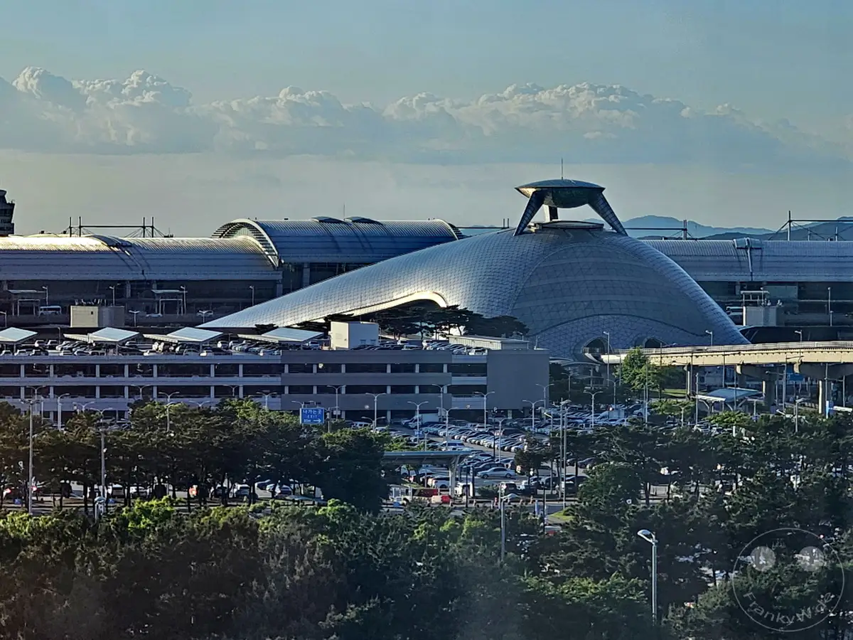 Der Internationale Flughafen Incheon in Südkorea besticht durch seine futuristische Architektur, die sich unter einem weiten Himmel mit Schäfchenwolken abzeichnet. Eine weitläufige Parkplatzanlage und Bäume säumen die beeindruckende Gebäudestruktur. Die Szene vermittelt einen Eindruck von Weite und moderner Mobilität im asiatischen Raum.