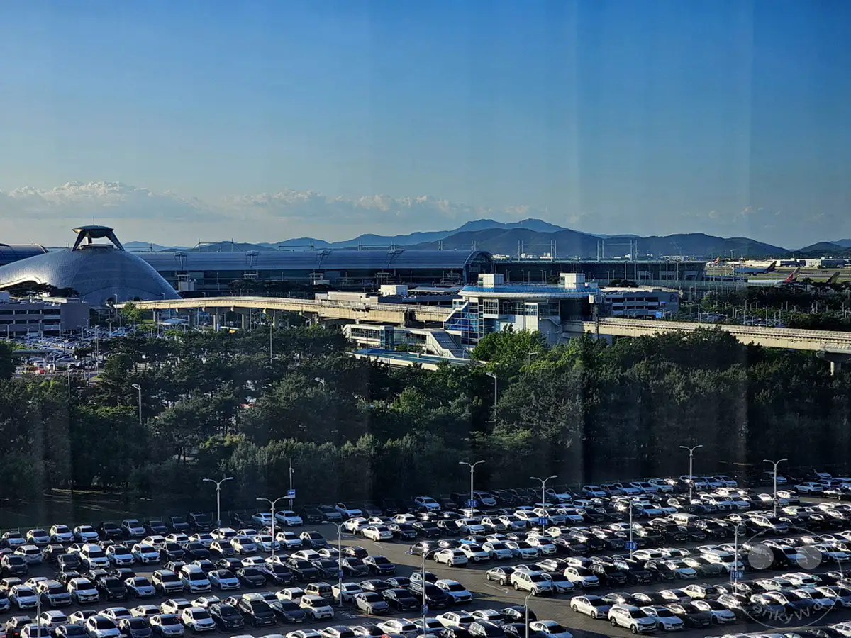 Eine Panoramaaufnahme des Incheon International Airport in Südkorea mit Blick auf mehrere Terminals, eine weitläufige Parkfläche voller Autos und im Hintergrund eine Bergkette. Das klare Wetter und das warme Licht deuten auf einen sonnigen Tag hin, der die moderne Infrastruktur des Flughafens gut zur Geltung bringt. Zahlreiche Flugzeuge sind auf dem Rollfeld sichtbar, was die Betriebsamkeit des internationalen Drehkreuzes unterstreicht.