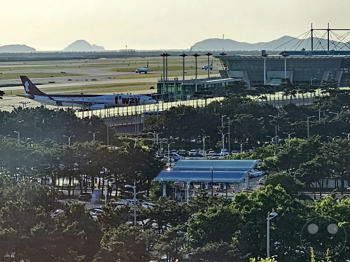 Ein Flugzeug der Marke T'way Air steht auf dem Rollfeld des Flughafens Incheon in der Nähe des Terminals. Im Hintergrund sind Berge in der sanften Morgendämmerung zu sehen, während Bäume und Parkplätze das Vordergrundbild dominieren. Die Szene vermittelt eine ruhige und geschäftige Atmosphäre eines internationalen Drehkreuzes.