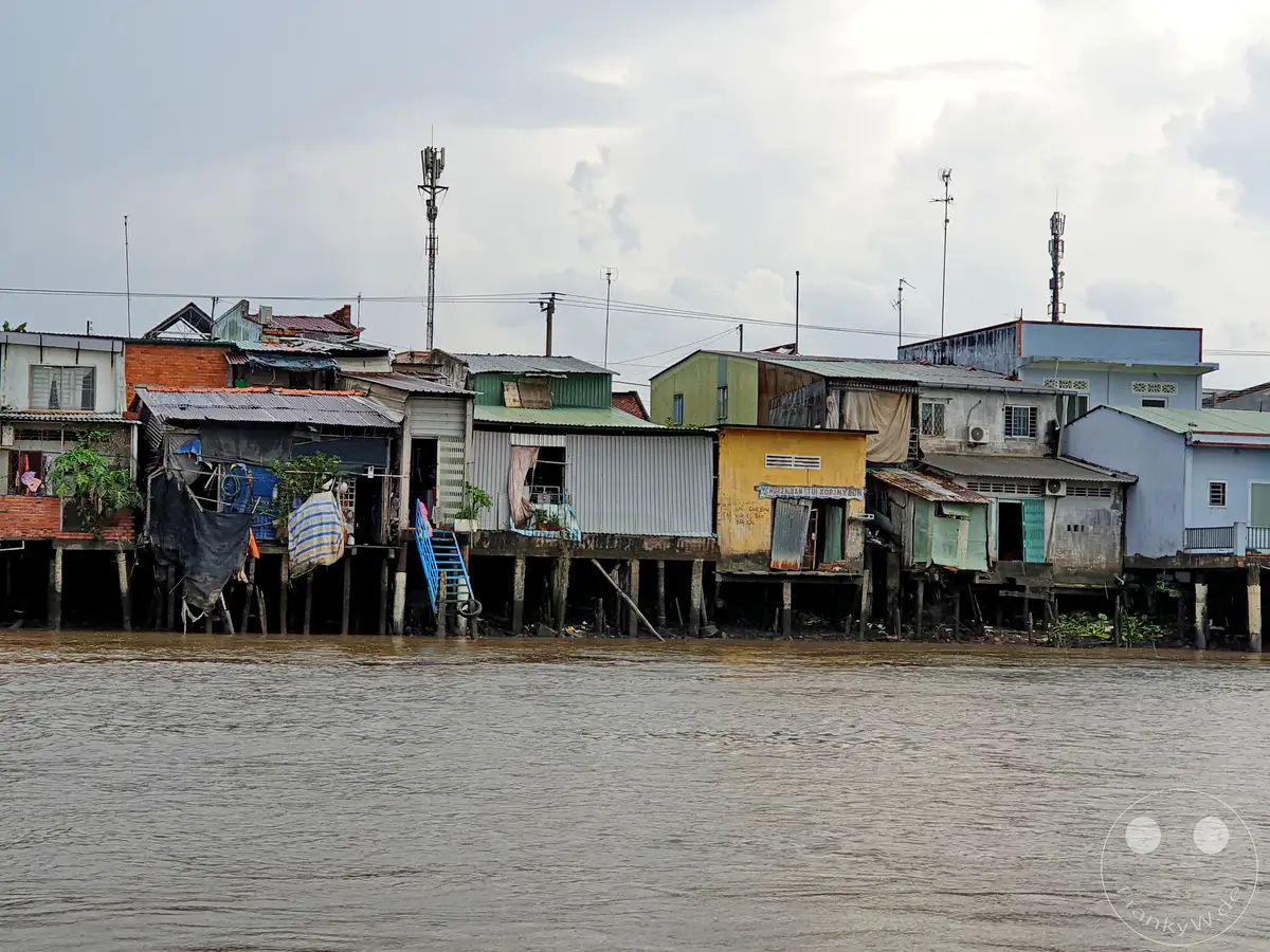 Die unsichere Existenz von Pfahlbauten am Ufer des Mekong in Vietnam wird durch die rustikalen und improvisierten Strukturen betont. Eine dichte, bunte Siedlung mit unterschiedlichen Gebäuden ragt über dem trüben Wasser empor und vermittelt eine Atmosphäre von Widerstandsfähigkeit angesichts widriger Umstände. Die Szene fängt das alltägliche Leben und die architektonische Vielfalt dieser Flussgemeinschaft ein.