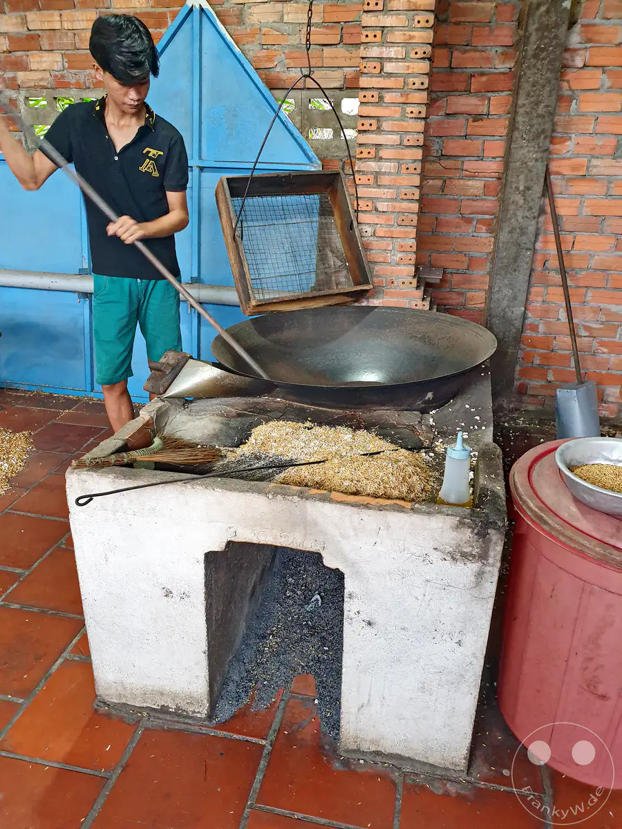 In Vietnam, am Mekong-Delta, wird in einer traditionellen Manufaktur Reispuffer hergestellt. Ein junger Mann rührt die Reiskörner über einem offenen Feuer in einem großen Wok, um sie zu puffen. Die Szene strahlt eine rustikale und authentische Atmosphäre aus, typisch für ländliche Handwerksbetriebe.
