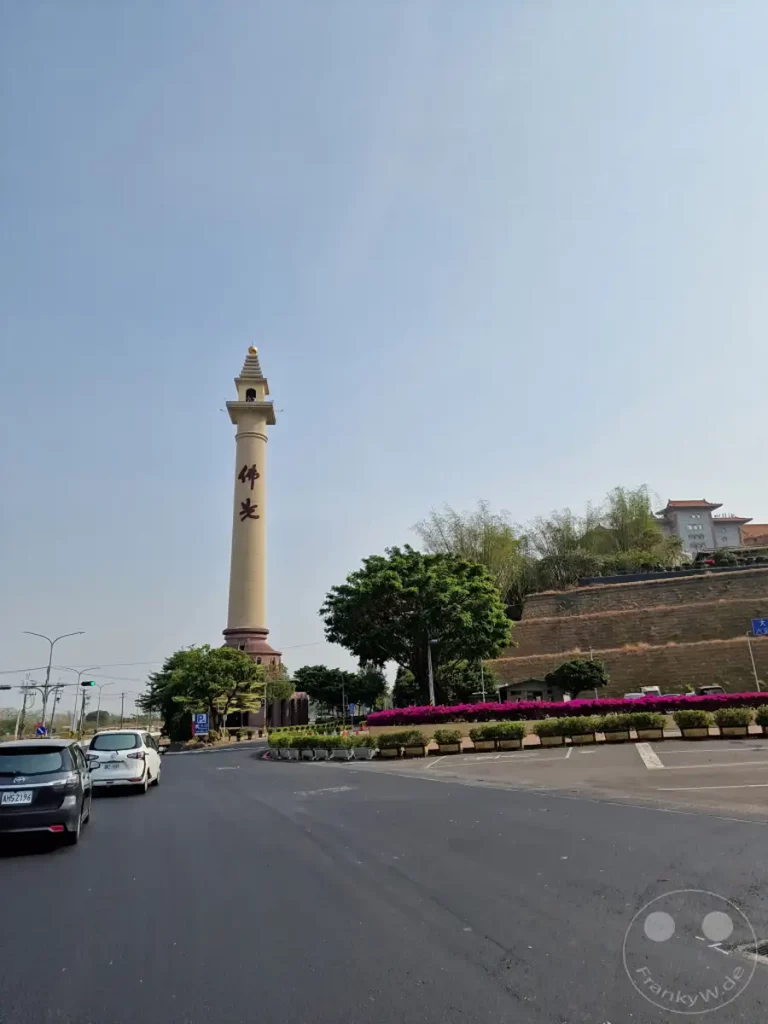 Ein hoher, beiger Turm mit chinesischen Schriftzeichen und einer goldenen Spitze dominiert die Szene vor einem klaren blauen Himmel. Im Hintergrund erstreckt sich eine terrassierte Hügelwand, geschmückt mit üppigen grünen Bäumen und leuchtend pinken Blumenbeeten, die die friedliche Atmosphäre der buddhistischen Stätte unterstreichen. Autos passieren eine Straße, die zu dem beeindruckenden Bauwerk führt und das alltägliche Leben mit der spirituellen Umgebung verbindet.