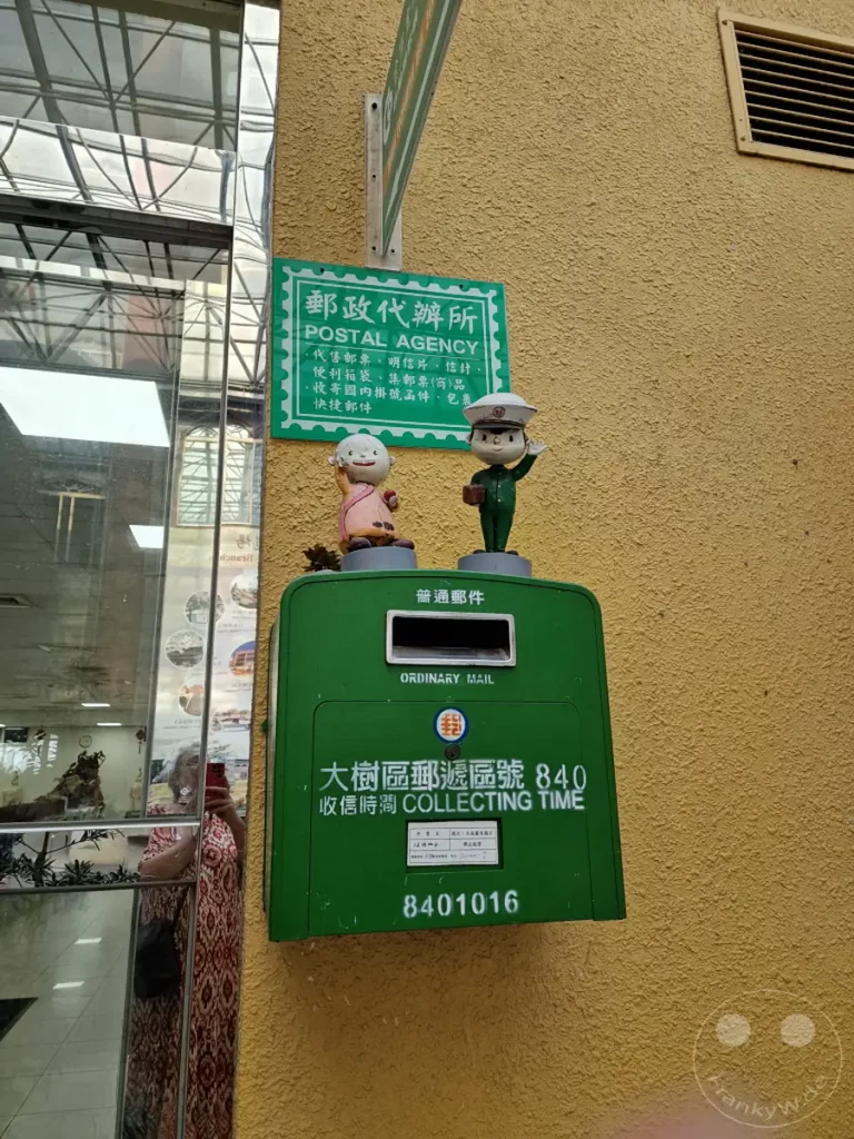 Vor dem Fo Guang Shan Kloster in Kaohsiung, Taiwan, steht ein grüner Briefkasten, der als Postschalter dient. Zwei niedliche Figuren, die einen Postboten und eine ältere Dame darstellen, schmücken die Vorderseite des Briefkastens und verleihen dem Ort eine freundliche Atmosphäre. Die Einrichtung ist funktional und dennoch mit charmanten Details versehen, die typisch für die Kultur der Region sind.