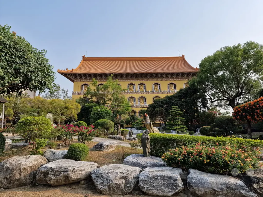 Die majestätische Fassade des Fo Guang Shan Monastery in Kaohsiung, Taiwan, ragt über einer üppigen Gartenanlage auf. Zahlreiche Buddha-Statuen sind in den Fenstern des gelben Gebäudes zu erkennen, während skulpturale Elemente und blühende Sträucher die vordere Szenerie beleben. Die friedliche Atmosphäre des buddhistischen Zentrums wird durch die klare Himmelskulisse und die gepflegte Landschaft unterstrichen.