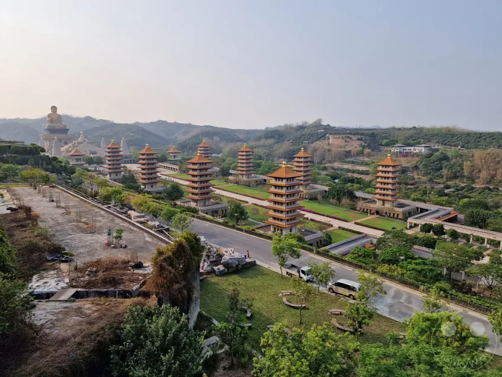 Ein weiter Blick auf den Fo Guang Shan Buddha Museum Komplex in Kaohsiung, Taiwan, enthüllt mehrere traditionelle Pagoden, die sich harmonisch in die hügelige Landschaft einfügen. Im Hintergrund erhebt sich eine monumentale Buddha-Statue vor einem hellen Himmel, was eine friedliche und erhabene Atmosphäre schafft. Die weitläufige Anlage mit ihren gepflegten Grünflächen und architektonischen Meisterwerken lädt zur Besinnung und Erkundung ein.