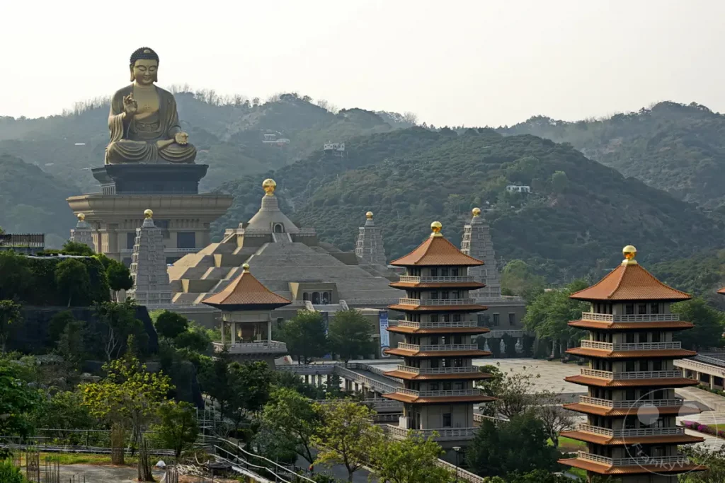Die monumentale Buddha-Statue thront über einem weitläufigen Tempelkomplex in Taiwan. Mehrere mehrstöckige Pagoden mit orangefarbenen Dächern und goldenen Spitzen sind im Vordergrund und Mittelgrund zu sehen. Die Szenerie strahlt eine tiefe Ruhe und Erhabenheit aus, eingebettet in die sanften Hügel des Landes.
